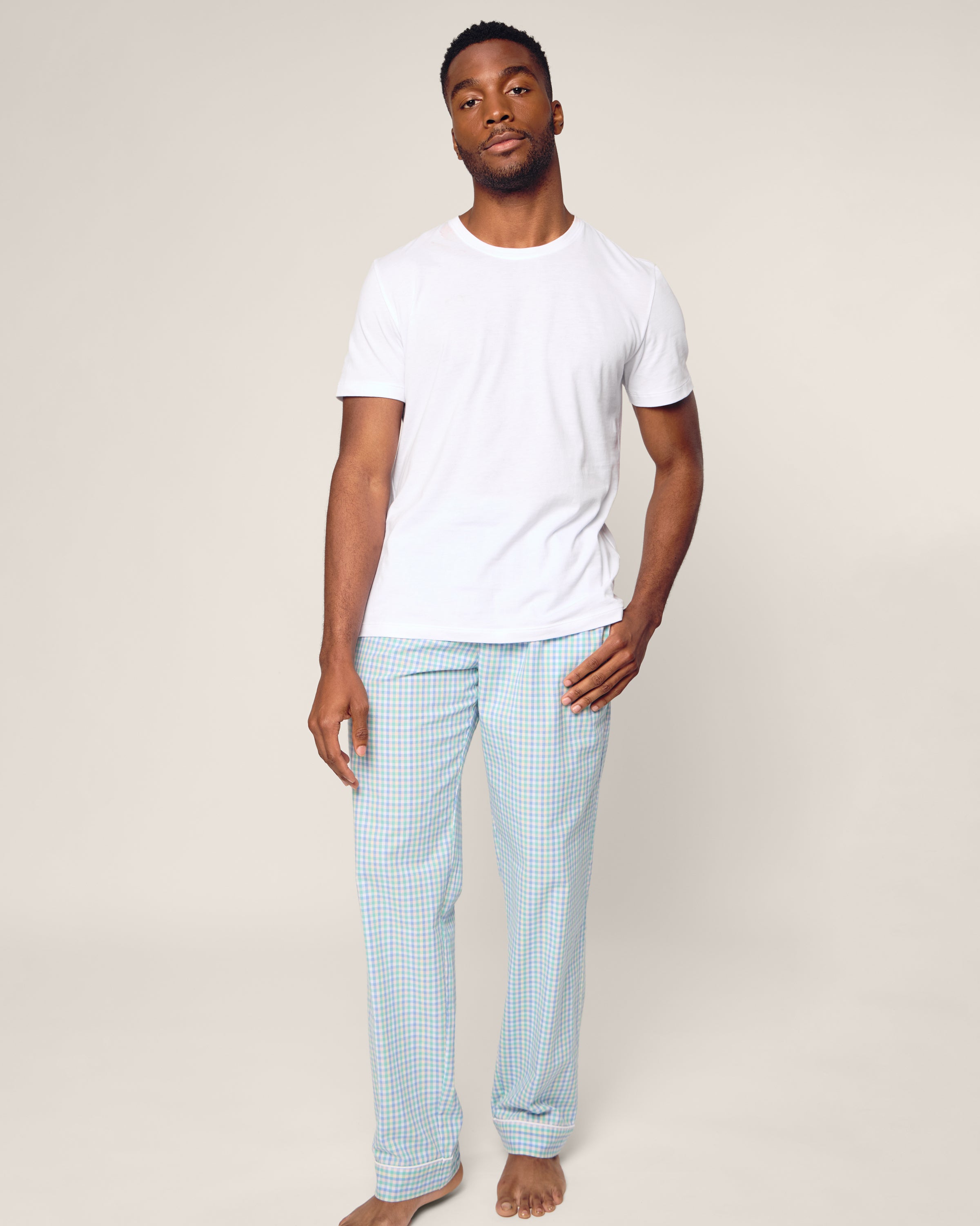 A person stands barefoot against a plain background, wearing luxurious sleepwear by Petite Plume, featuring a white T-shirt and Mens Twill Pajama Pants in Spring Gingham, crafted from the finest quality cotton. They look directly at the camera with a neutral expression.