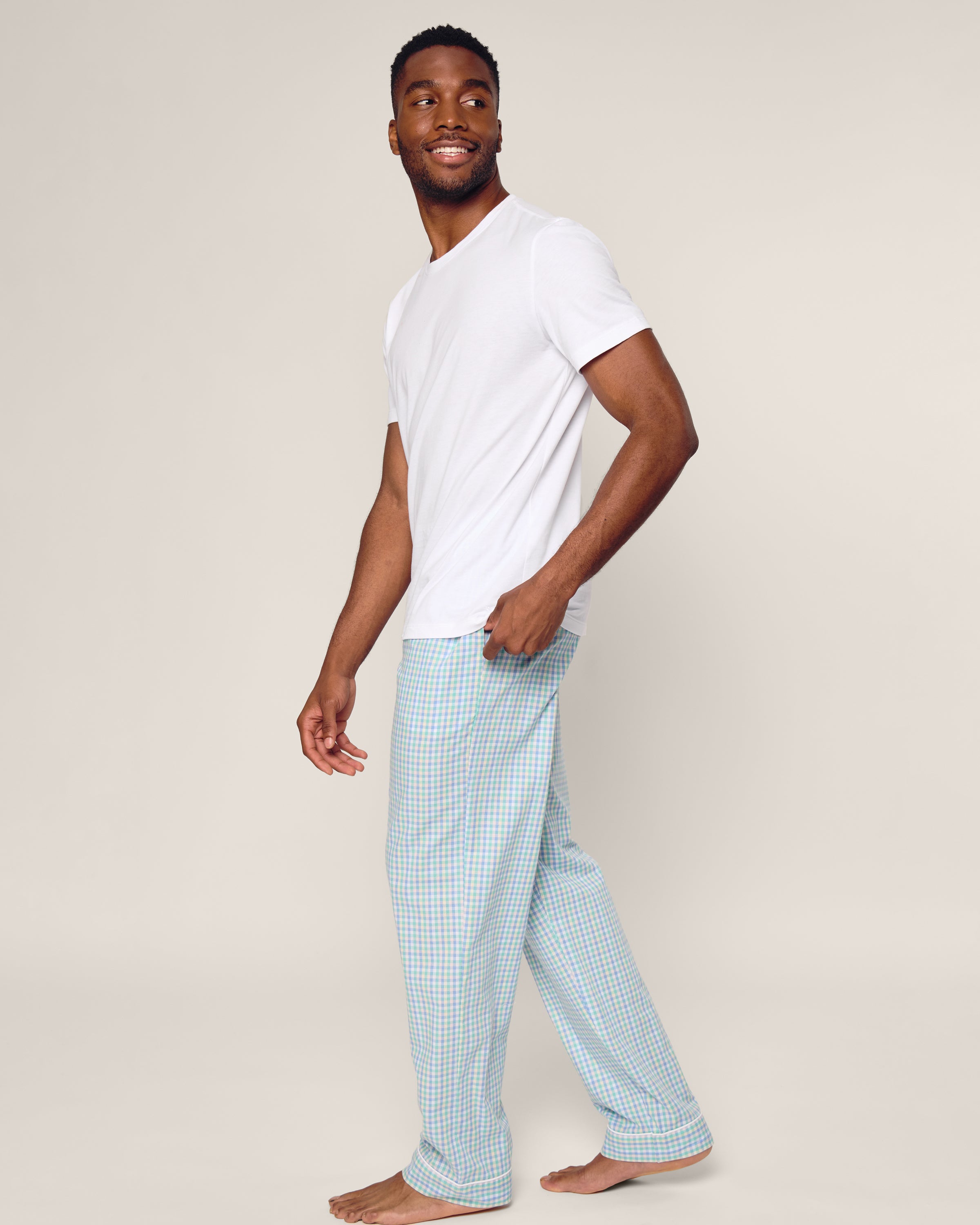 A barefoot person smiles while looking to the side, wearing a white T-shirt and Petite Plumes Mens Twill Pajama Pants in Spring Gingham made of finest cotton. The background is plain beige.