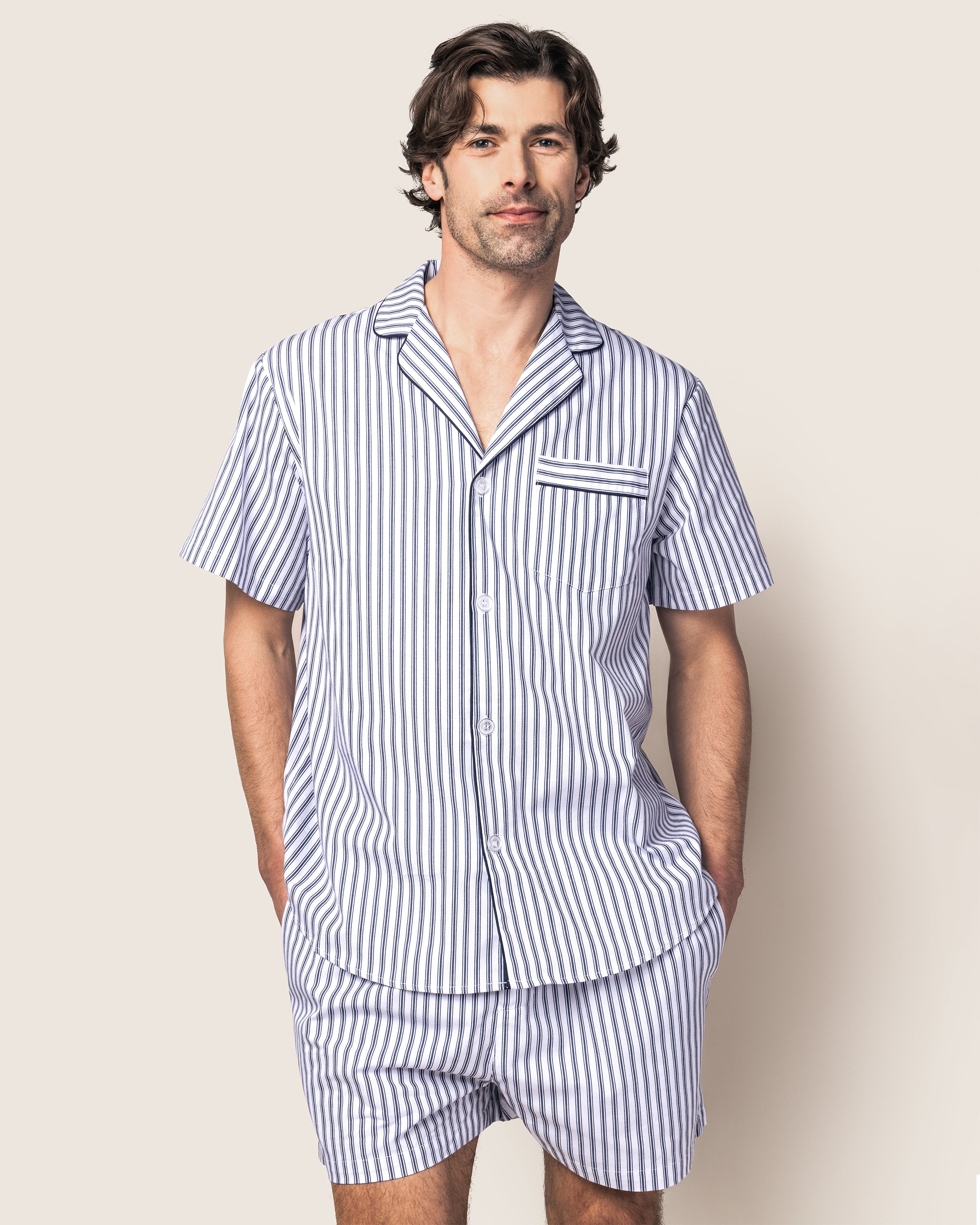 A person stands smiling slightly with hands in pockets, wearing the Petite Plume Mens Twill Pajama Short Set in Navy French Ticking against a plain beige background.