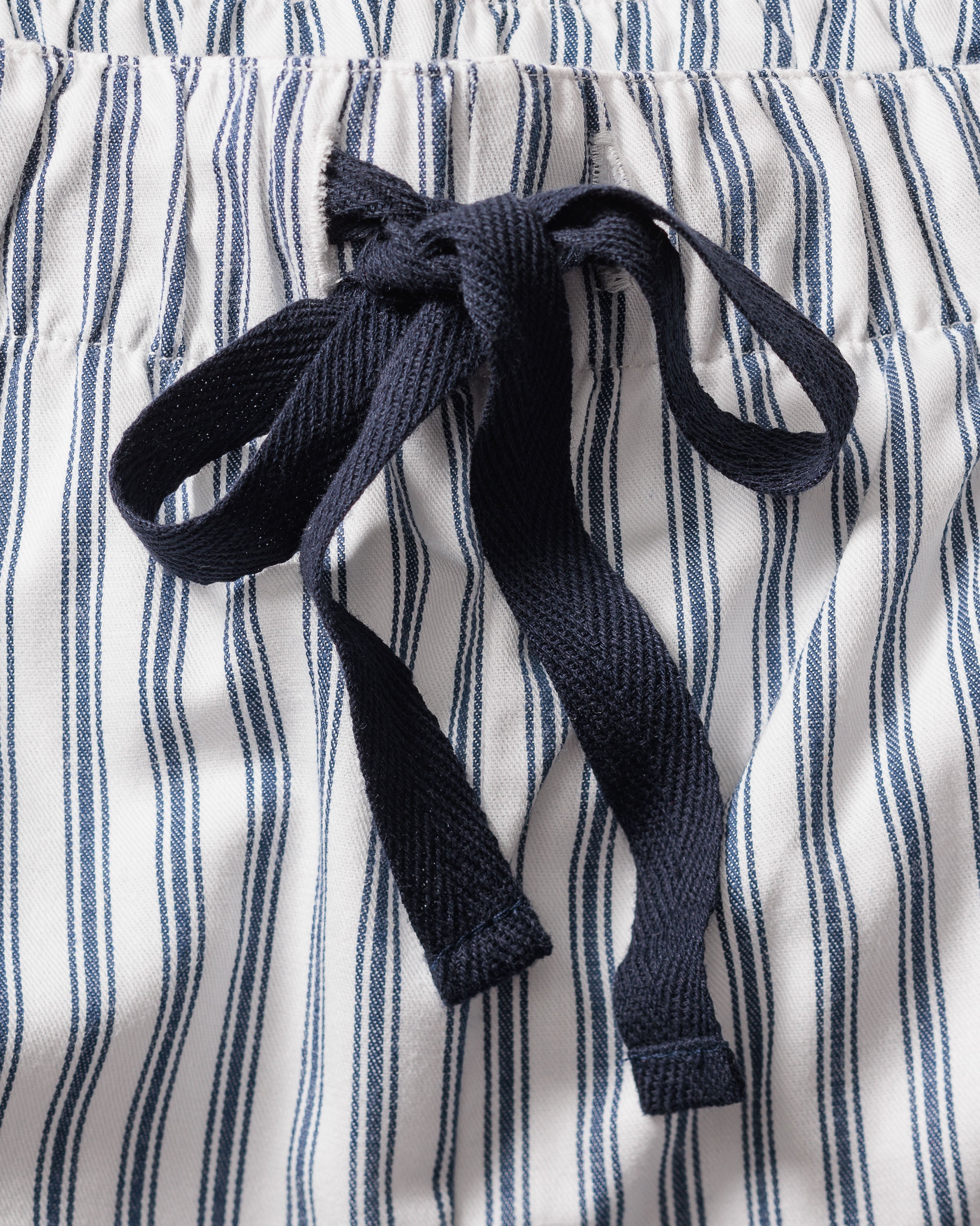 A close-up of the Mens Twill Pajama Short Set in Navy French Ticking by Petite Plume shows cotton shorts with blue and white vertical stripes, featuring an elastic waistband and black drawstring, ideal for relaxation.