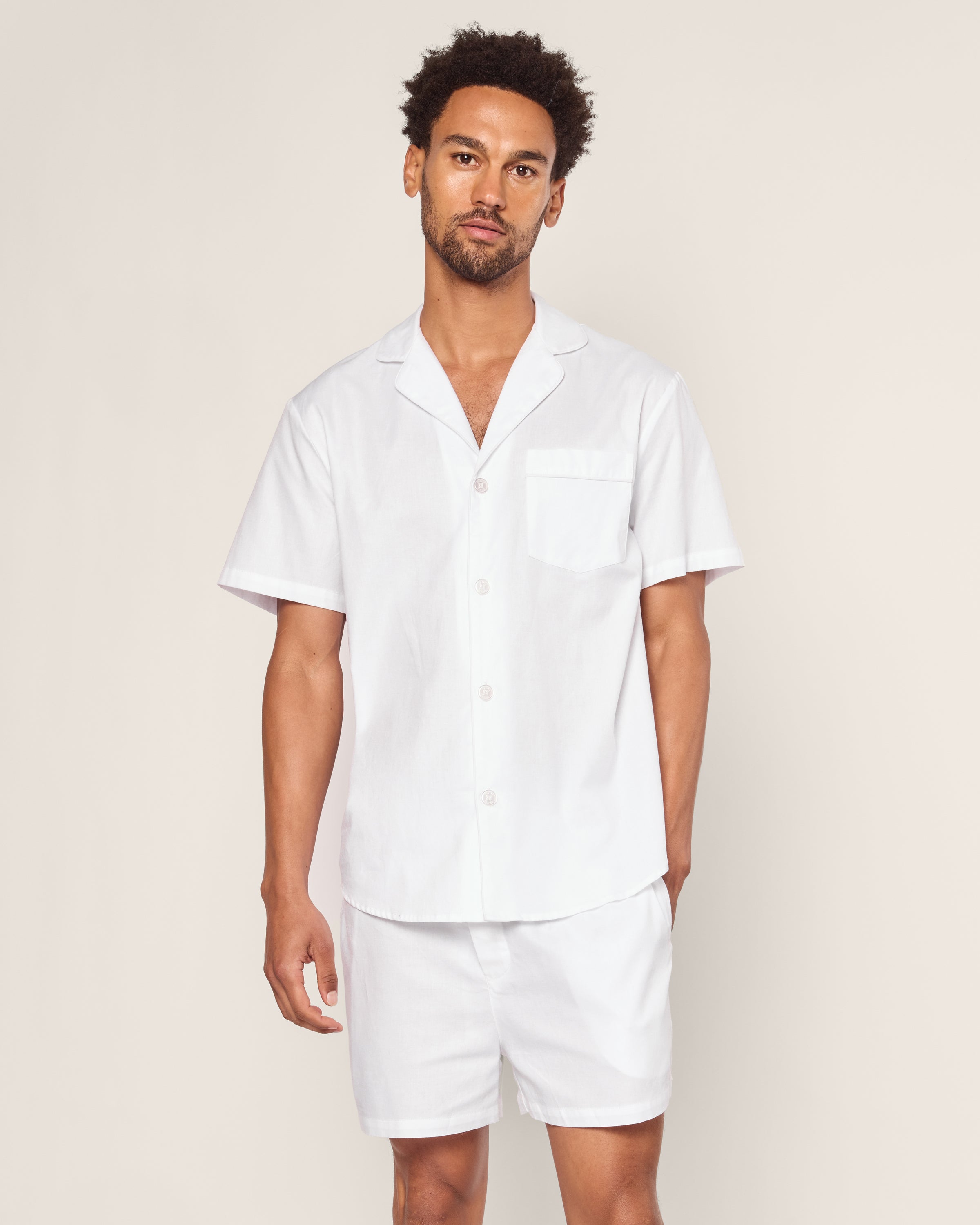 A man stands against a plain backdrop wearing the Mens Twill Pajama Short Set in white with white piping by Petite Plume, made from fine cotton. The short-sleeve button-up shirt, featuring a left pocket, complements his curly hair and beard with its crisp and comfy style.