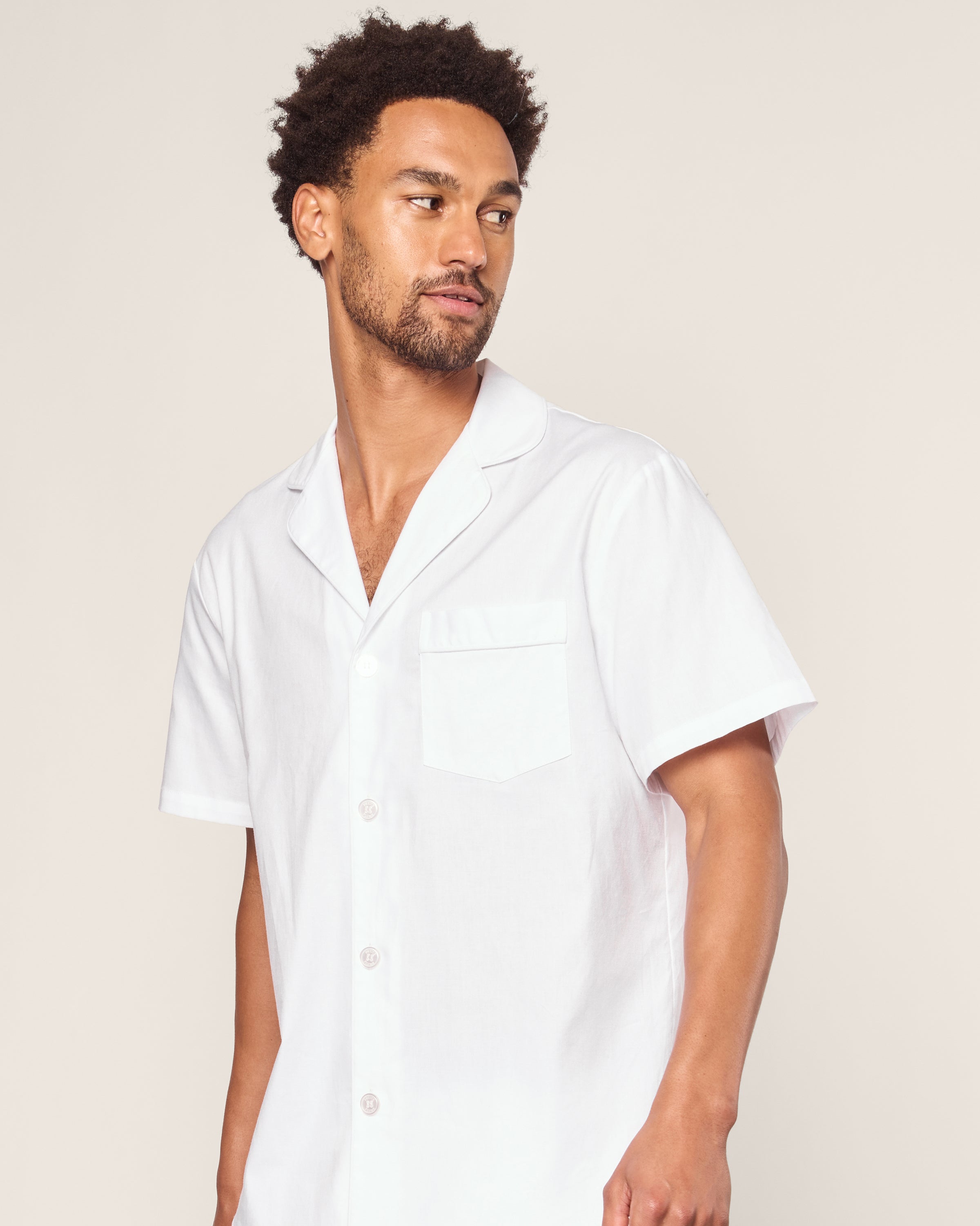 A man with short curly hair and a beard looks to the side, wearing Petite Plumes Mens Twill Pajama Short Set in white with white piping. The set includes a short-sleeve button-up shirt with a collar and chest pocket, made from the finest quality cotton. The background is plain and light-colored.