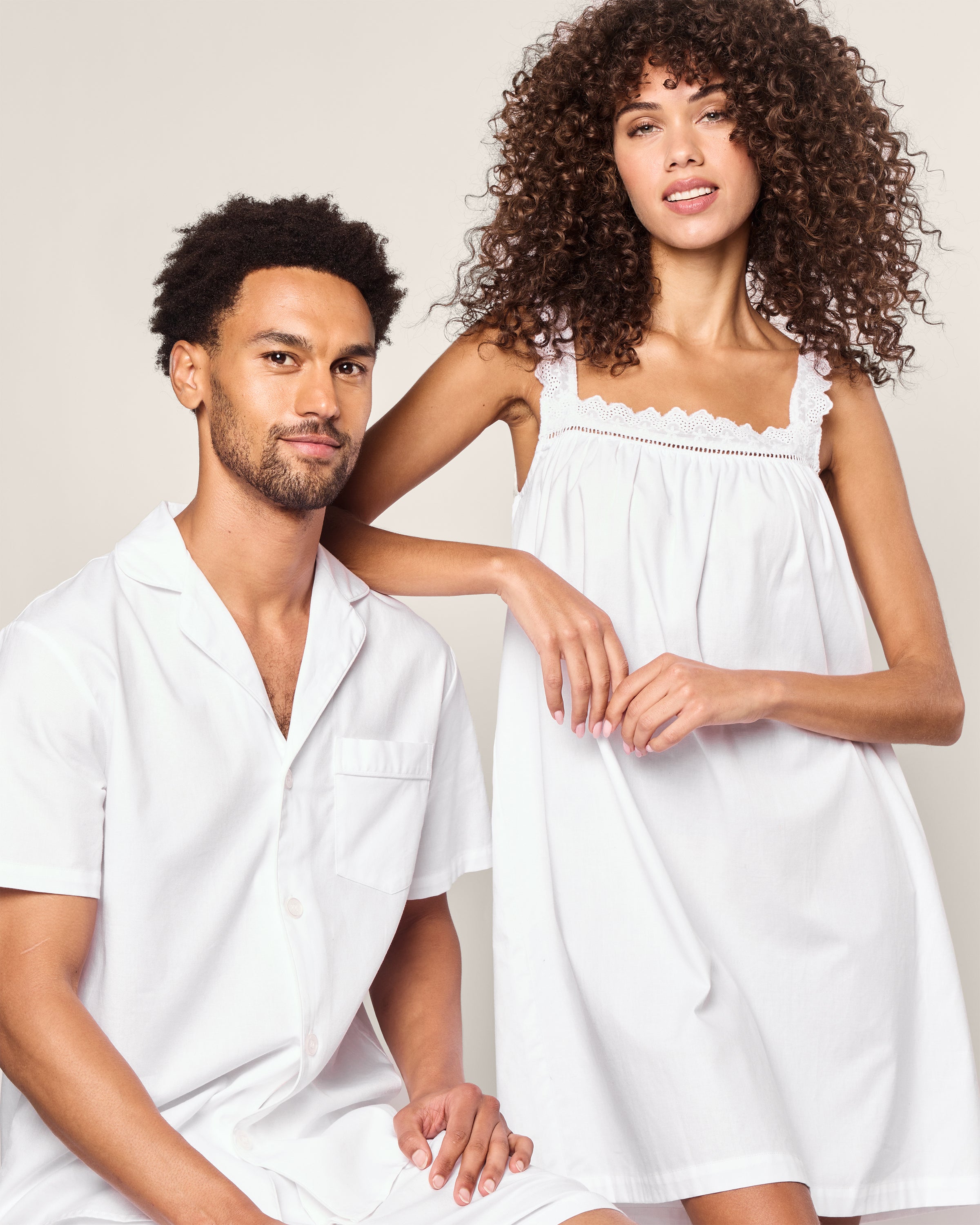 A couple showcases Petite Plume sleepwear. The man sits on a stool donning the Mens Twill Pajama Short Set White with White Piping, while the woman stands in a sleeveless nightdress with lace trim. Both outfits are crafted from high-quality cotton, as they smile against a plain backdrop.