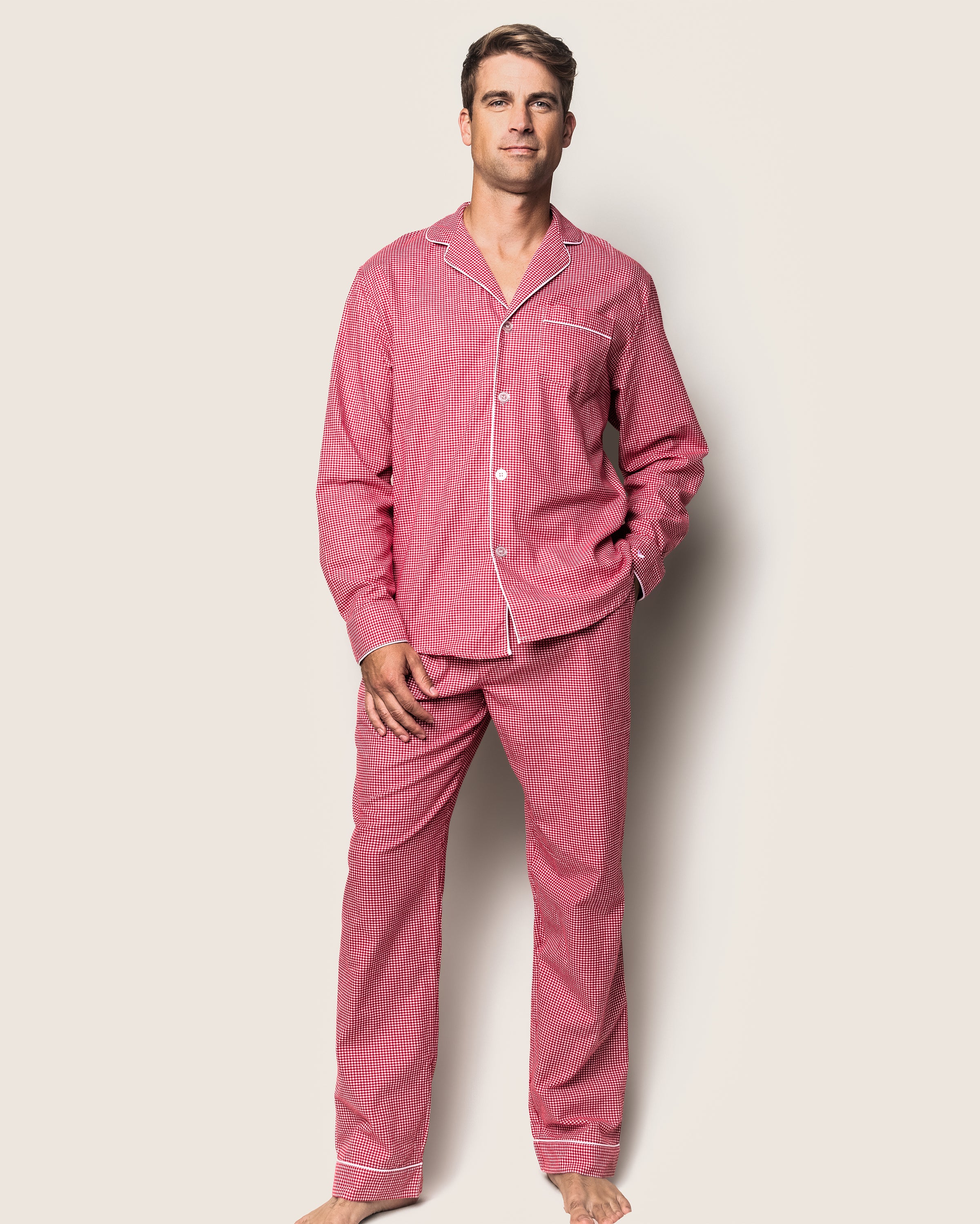 A person stands against a plain background wearing the Mens Flannel Pajama Set in Red Mini Gingham by Petite Plume, made from high-quality cotton. Their right hand is in the pants pocket as they appear relaxed and barefoot.