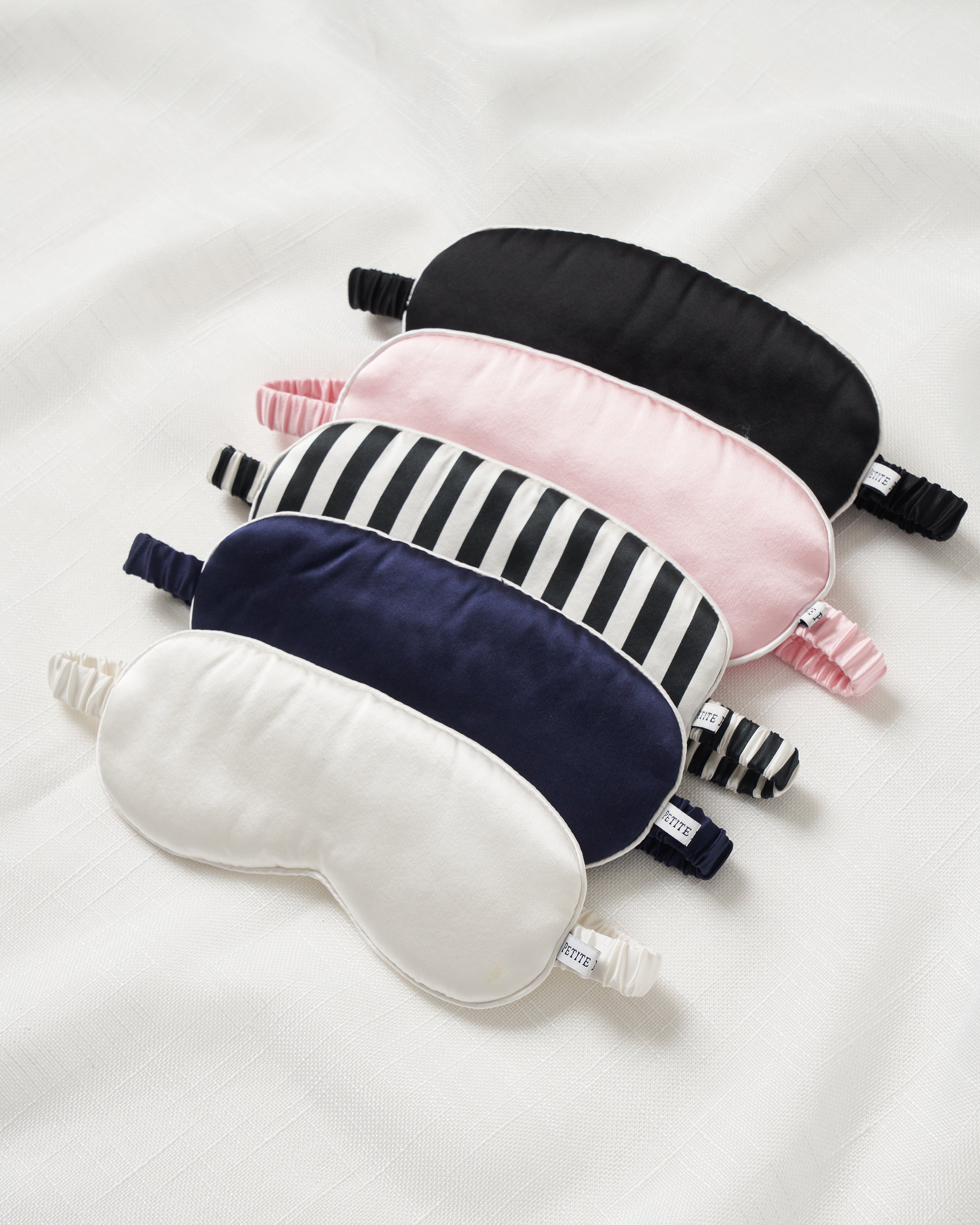 A neatly arranged selection of five top-quality silk sleep masks by Petite Plume, including designs in solid black, pink, navy (Adults Silk Sleep Mask in Navy), white, and black and white stripes, are displayed on a soft white fabric background.