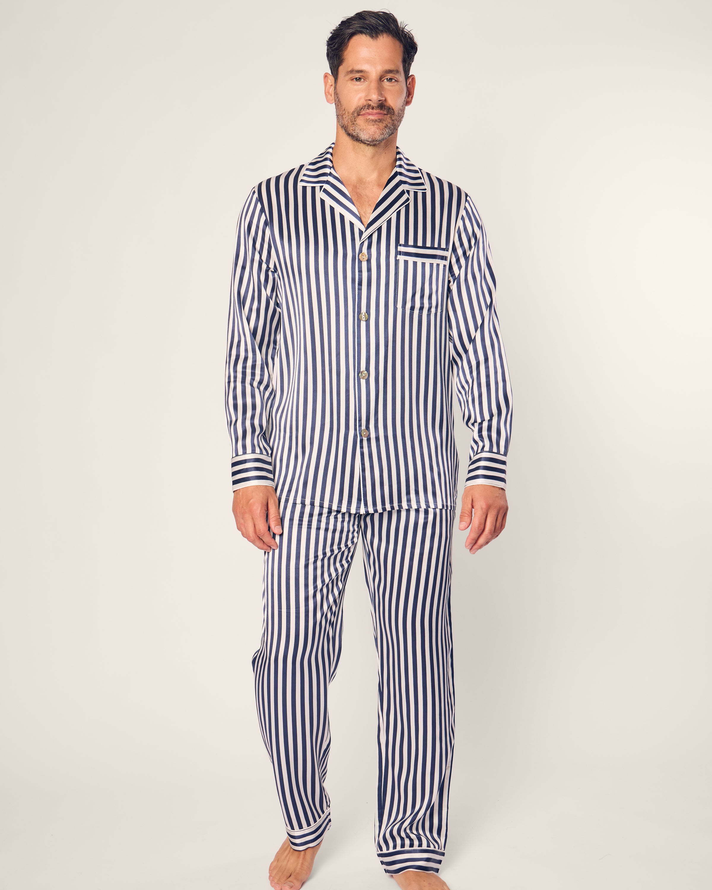 A man in a navy striped pajamas set from Petite Plume stands on a plain background. The ensemble, crafted from luxurious mulberry silk, includes a long-sleeved button-up shirt and pants. His beard and short hair add to his understated elegance.