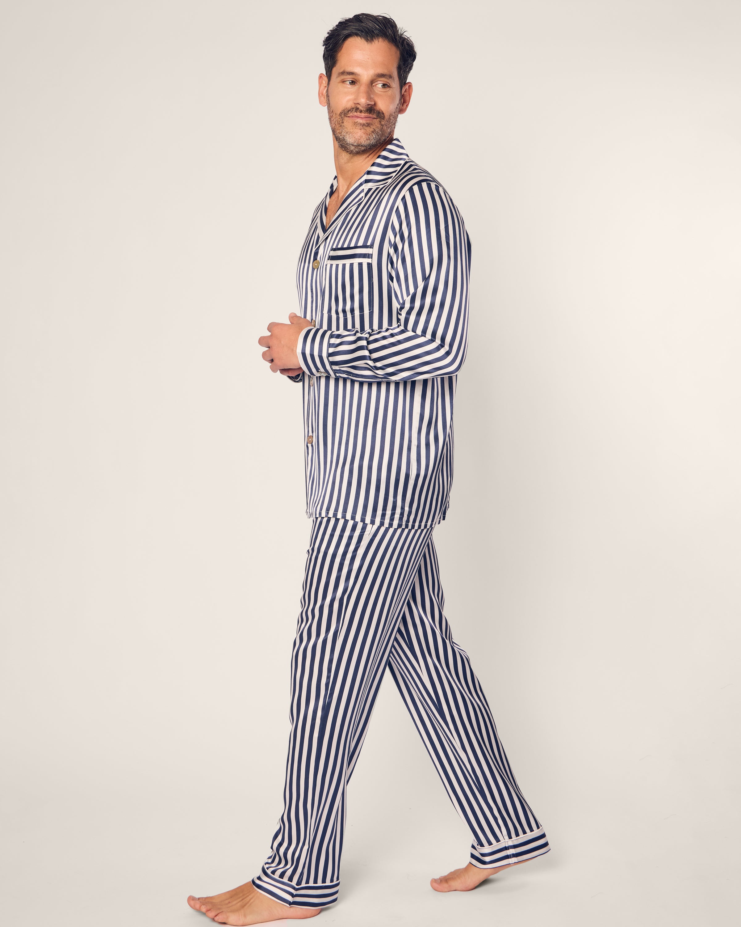 A man with a short beard and dark hair wears Petite Plume’s Mens Silk Pajama Set in Navy Bengal Stripe. Crafted from breathable mulberry silk, he walks barefoot on a plain background, glancing left with a slight smile.