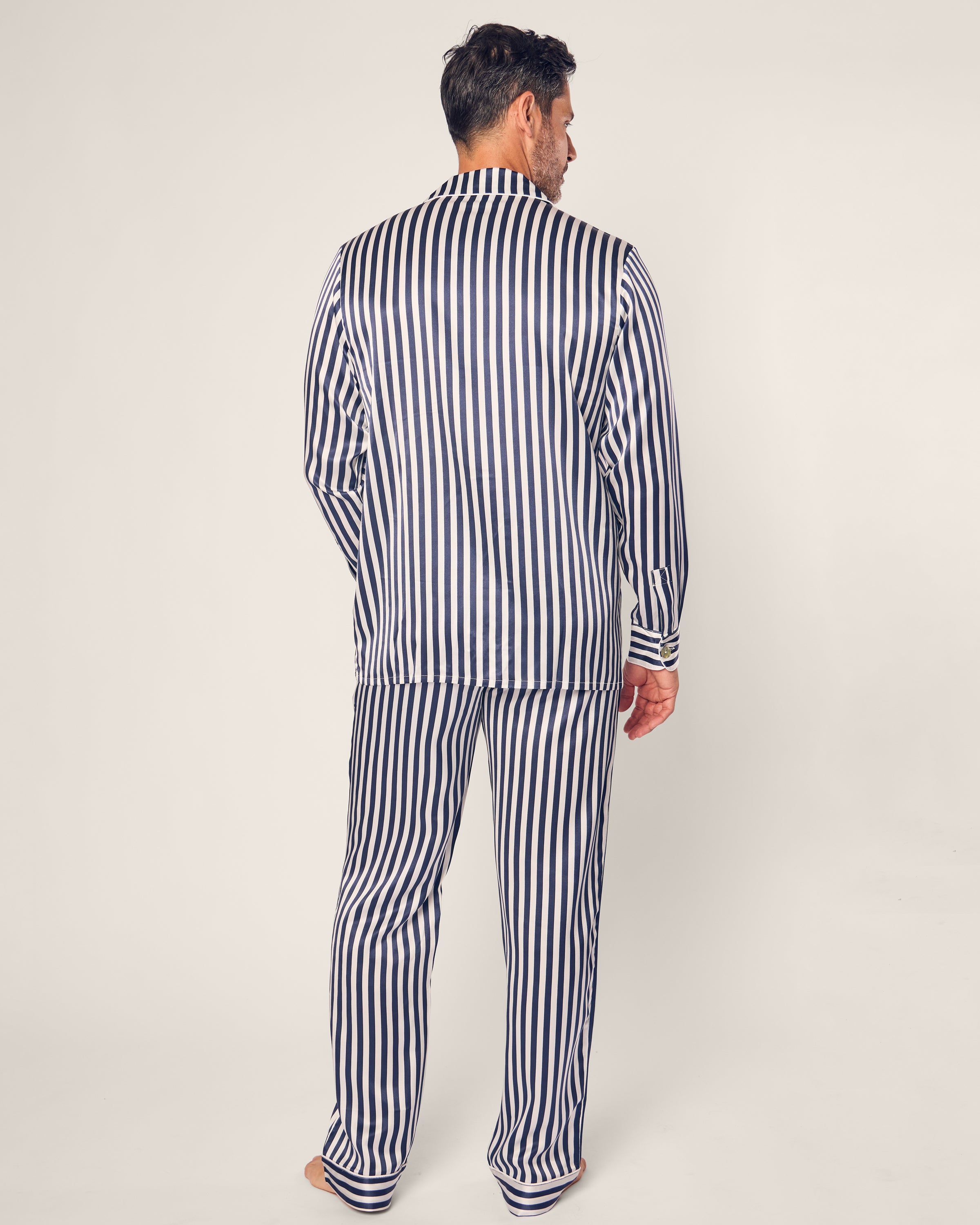 Barefoot against a plain background, a man wears Petite Plumes Mens Silk Pajama Set in Navy Bengal Stripe. Crafted from mulberry silk, the luxurious pajamas exude elegance as he stands facing away with his head slightly turned, highlighting the ensembles sleek design.