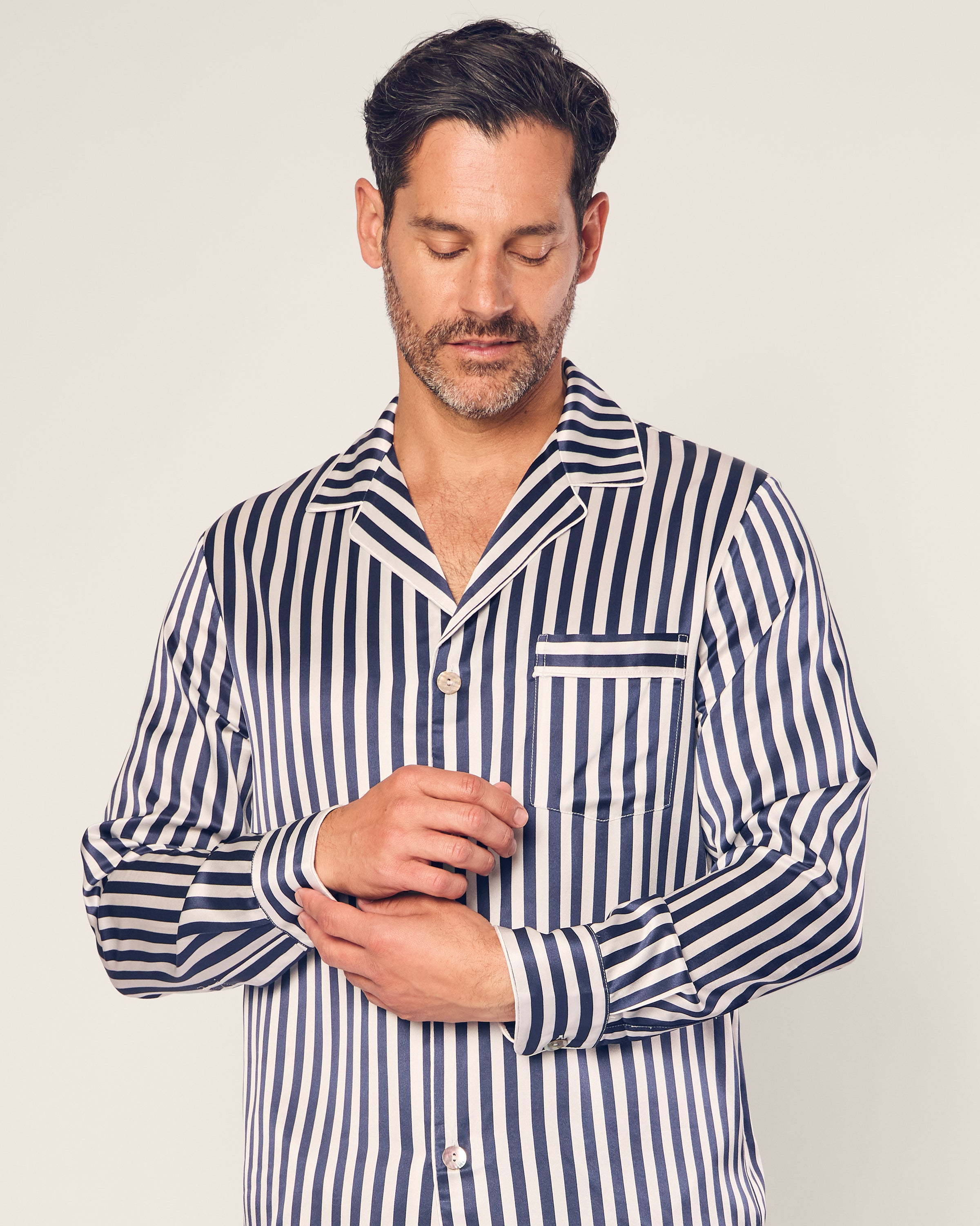 A man in Petite Plumes Mens Silk Pajama Set in Navy Bengal Stripe with breathable fabric adjusts one cuff, eyes closed. The top features blue and white vertical stripes and a chest pocket, epitomizing luxury sleepwear against a simple light backdrop.