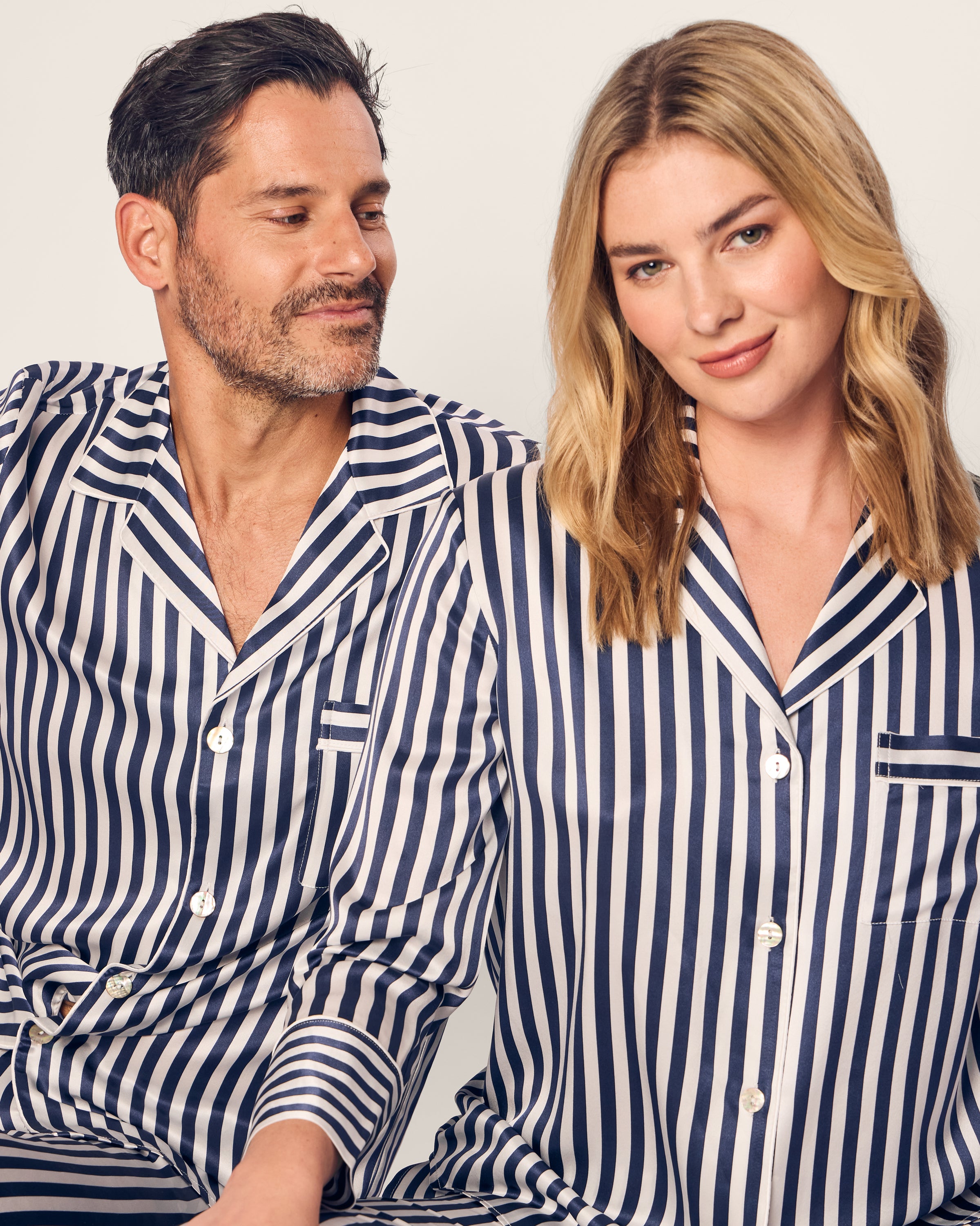 A man and woman sit side by side in matching Petite Plume Mens Silk Pajama Sets in Navy Bengal Stripe. The man, with short dark hair and a beard, smiles at the blonde-haired woman against a plain backdrop—embodying luxury sleepwear comfort.