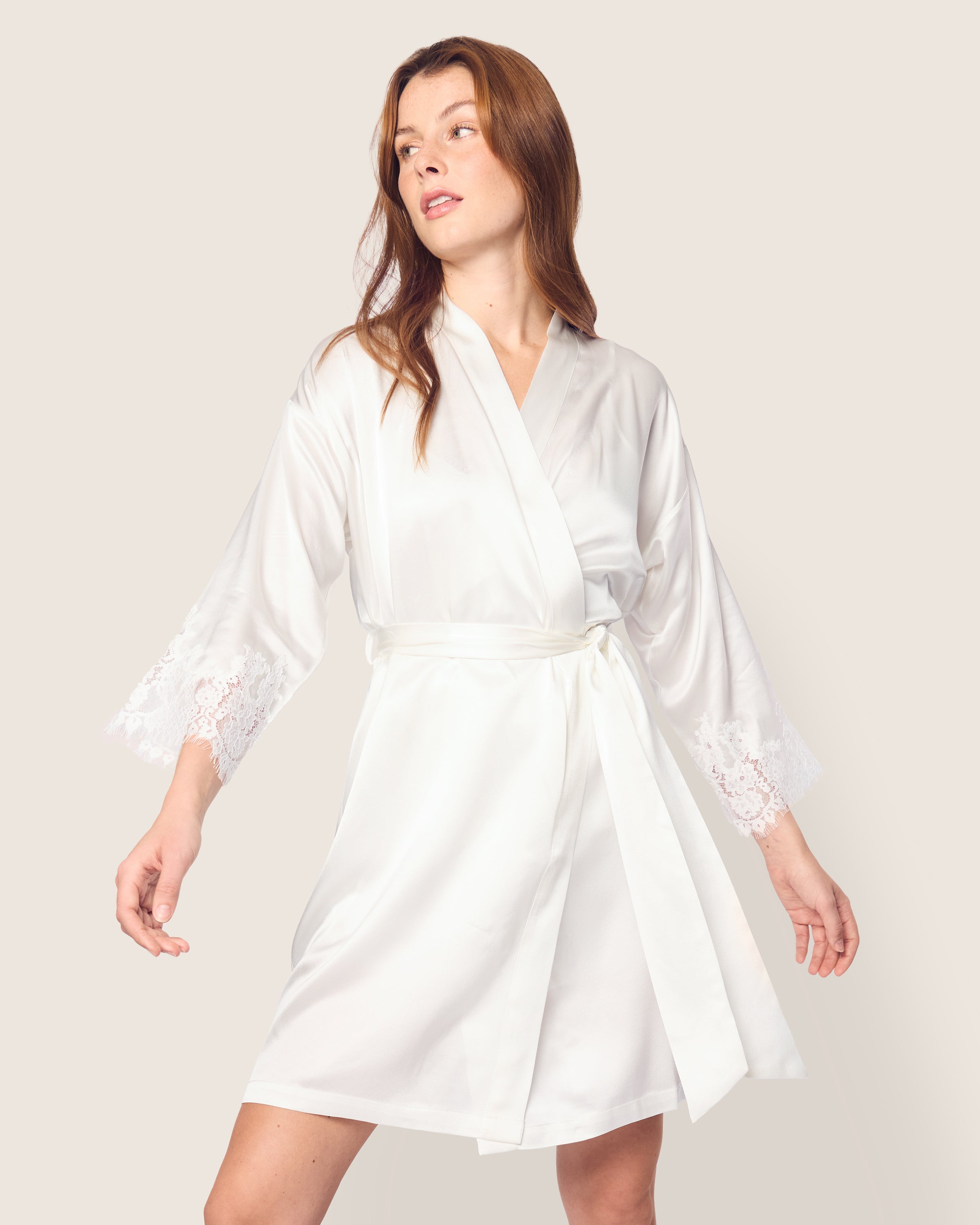 A person in a Womens Silk Lace Robe in White by Petite Plume stands against a plain background. With delicate lace sleeves and tied at the waist, this luxurious piece of sleepwear flows gently as they look to the side, one arm slightly extended.