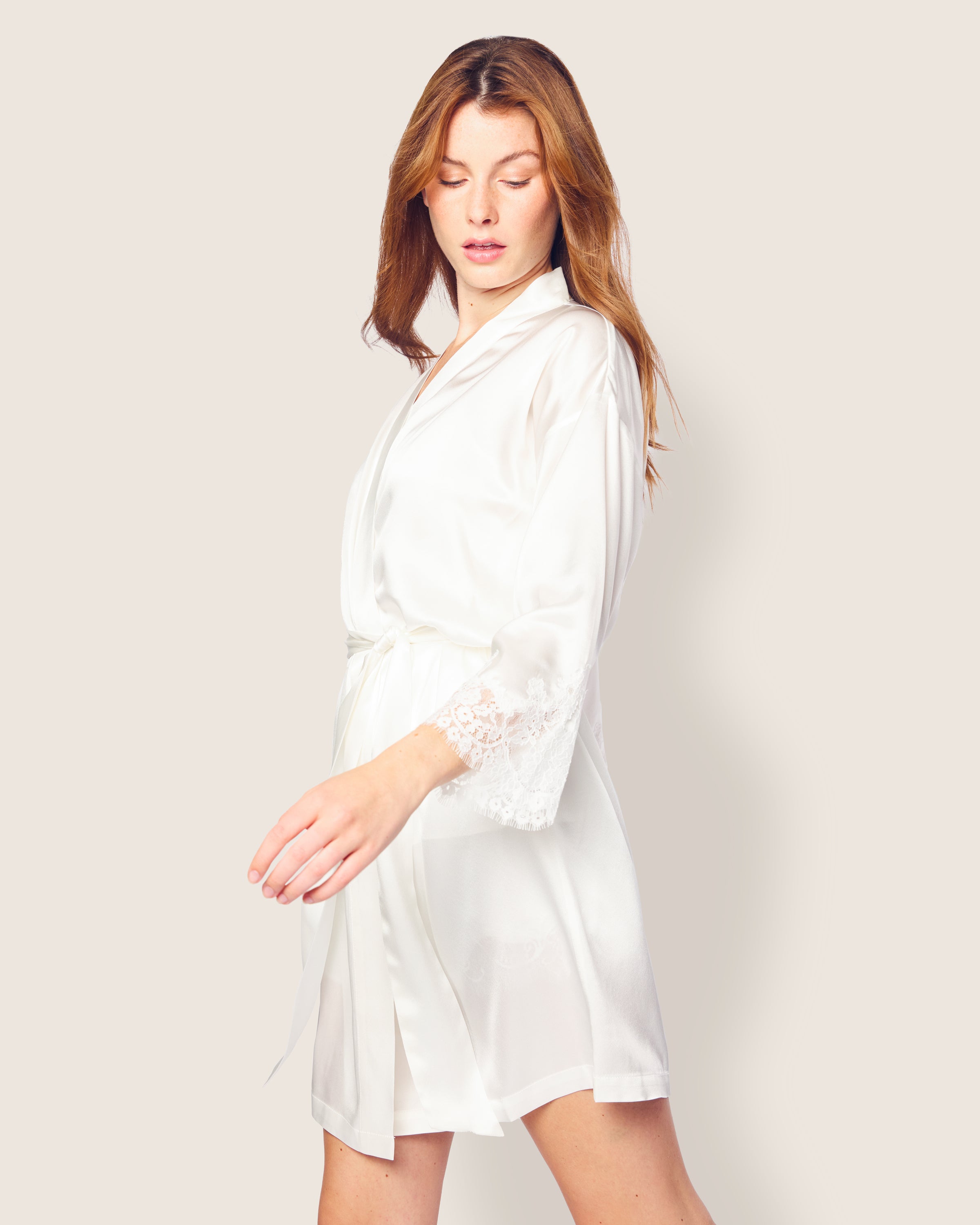 Wearing the luxurious Petite Plume Womens Silk Lace Robe in White, a person with long hair gracefully extends one arm to the side. The robe, made from mulberry silk with lace-trimmed sleeves, complements their look as they stand against a plain background looking slightly downwards.