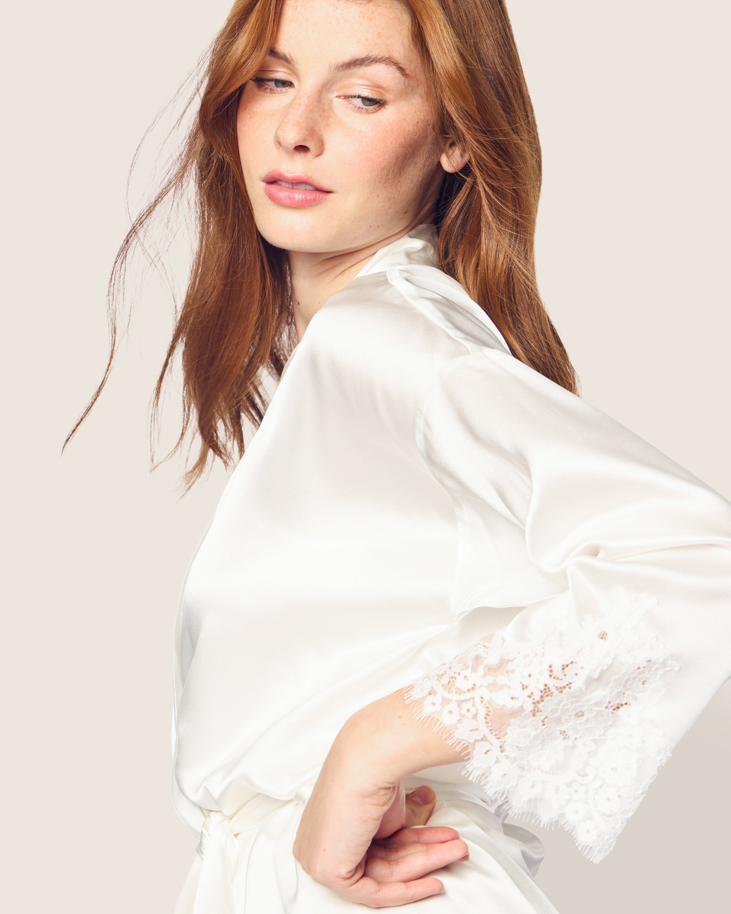 A red-haired woman models the Womens Silk Lace Robe in White by Petite Plume, showcasing its luxurious mulberry silk and lace sleeve details. She embraces its hypoallergenic comfort against a plain, light background with a relaxed expression.