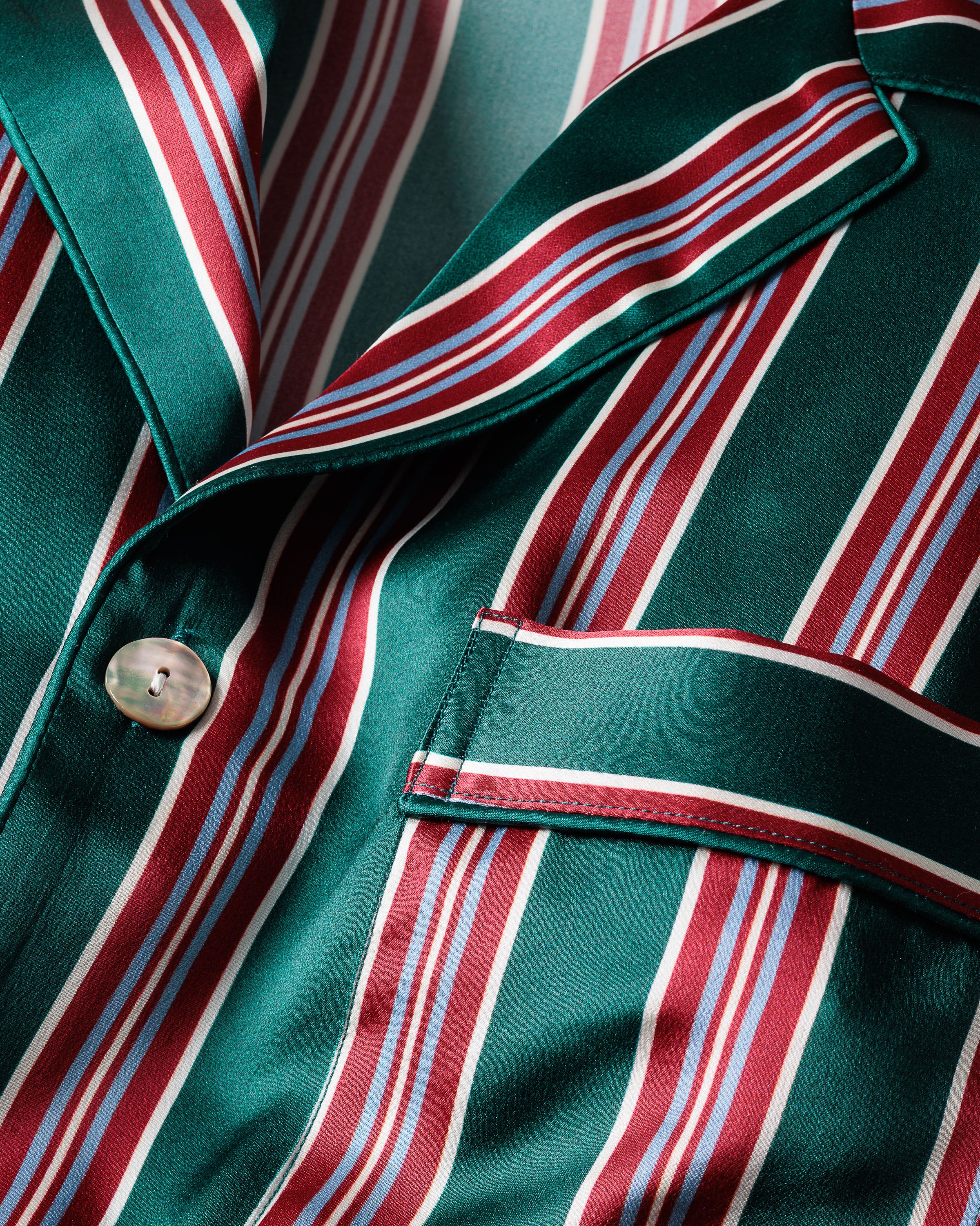 Close-up of a Womens Silk Nightshirt in Lancaster Stripe by Petite Plume, featuring green, red, and white vertical stripes. The shiny texture suggests Mulberry Silk, while a button and pocket indicate it could be part of elegant sleepwear or a stylish jacket or shirt.