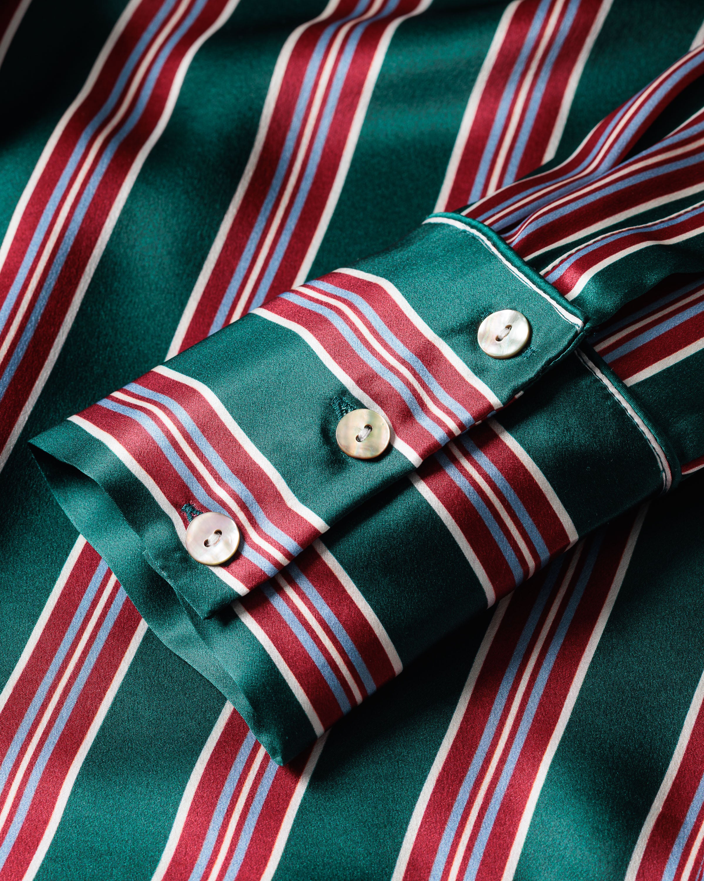 Close-up of a sleeve from the Womens Silk Nightshirt in Lancaster Stripe by Petite Plume, featuring green Mulberry Silk with vertical red, white, and light blue stripes. The cuff has two white buttons and a buttonhole, highlighting its smooth, luxurious pajama-like fabric.