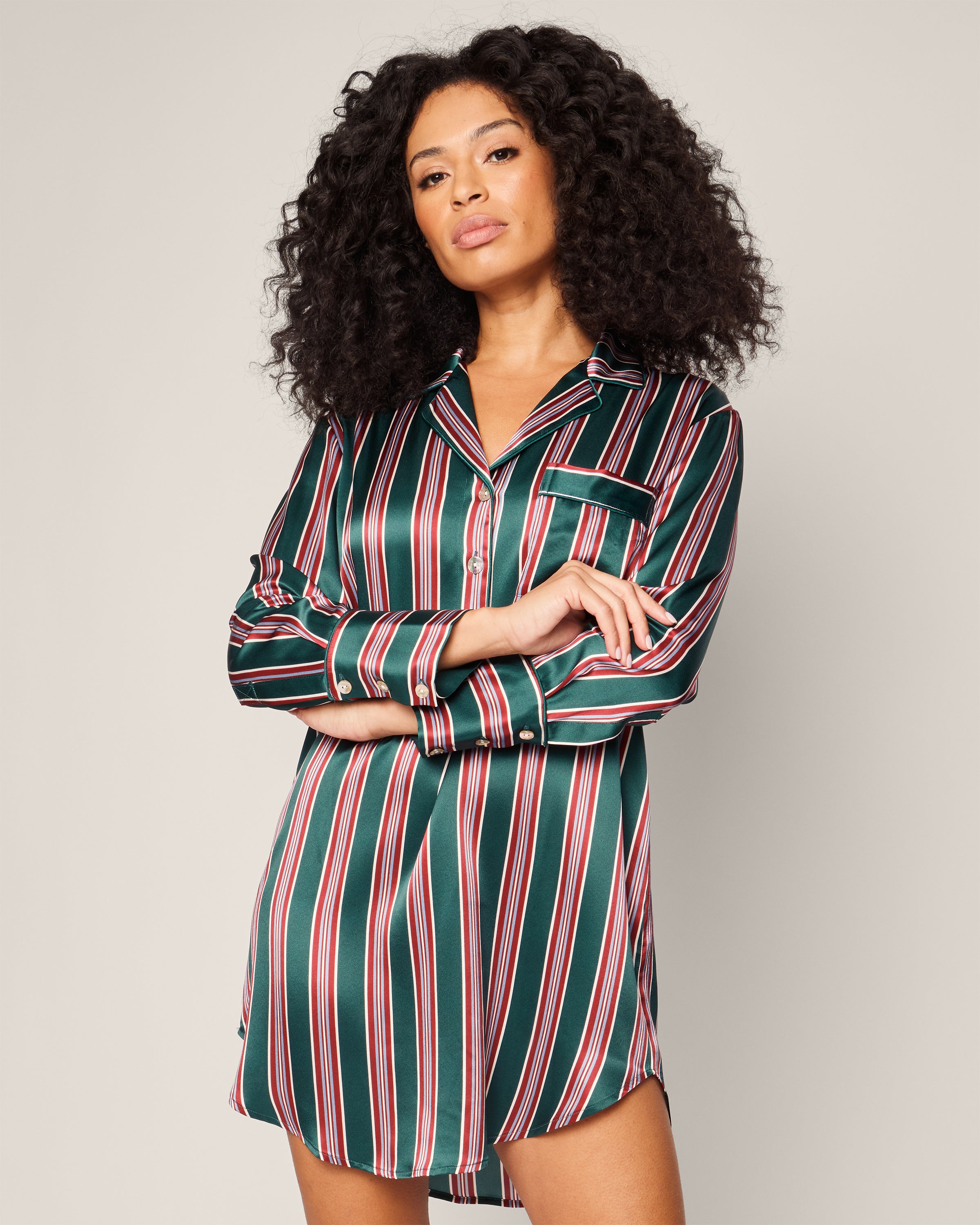 A person with long, curly hair confidently wears the Womens Silk Nightshirt in Lancaster Stripe by Petite Plume, featuring green, red, and white stripes. With arms crossed against a plain background, they exude luxurious sleepwear style.