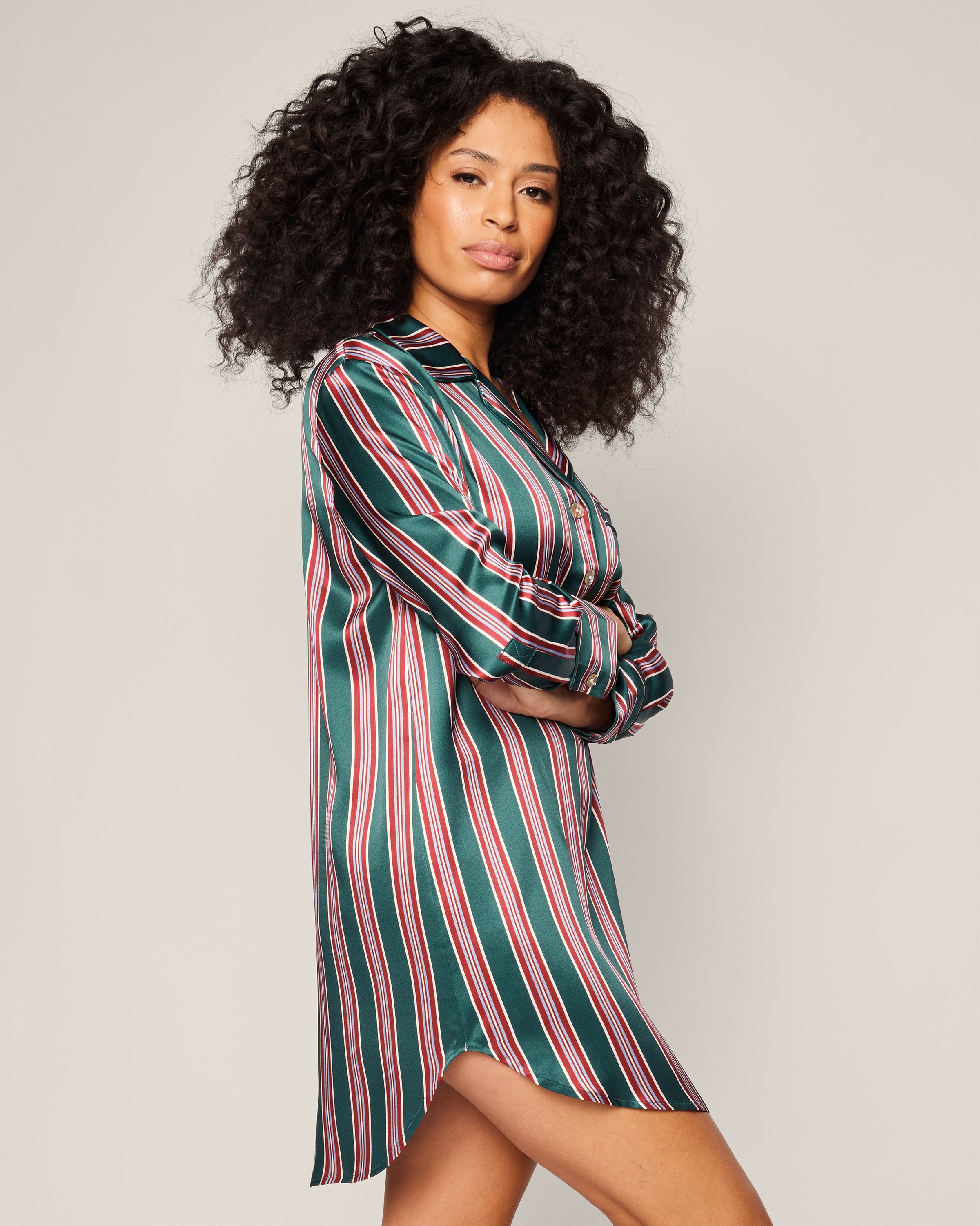 A person with long, curly hair stands confidently with crossed arms, wearing Petite Plumes Womens Silk Nightshirt in Lancaster Stripe. The green, red, and white stripes resemble a tailored dress against a plain light-colored background that highlights their striking presence.