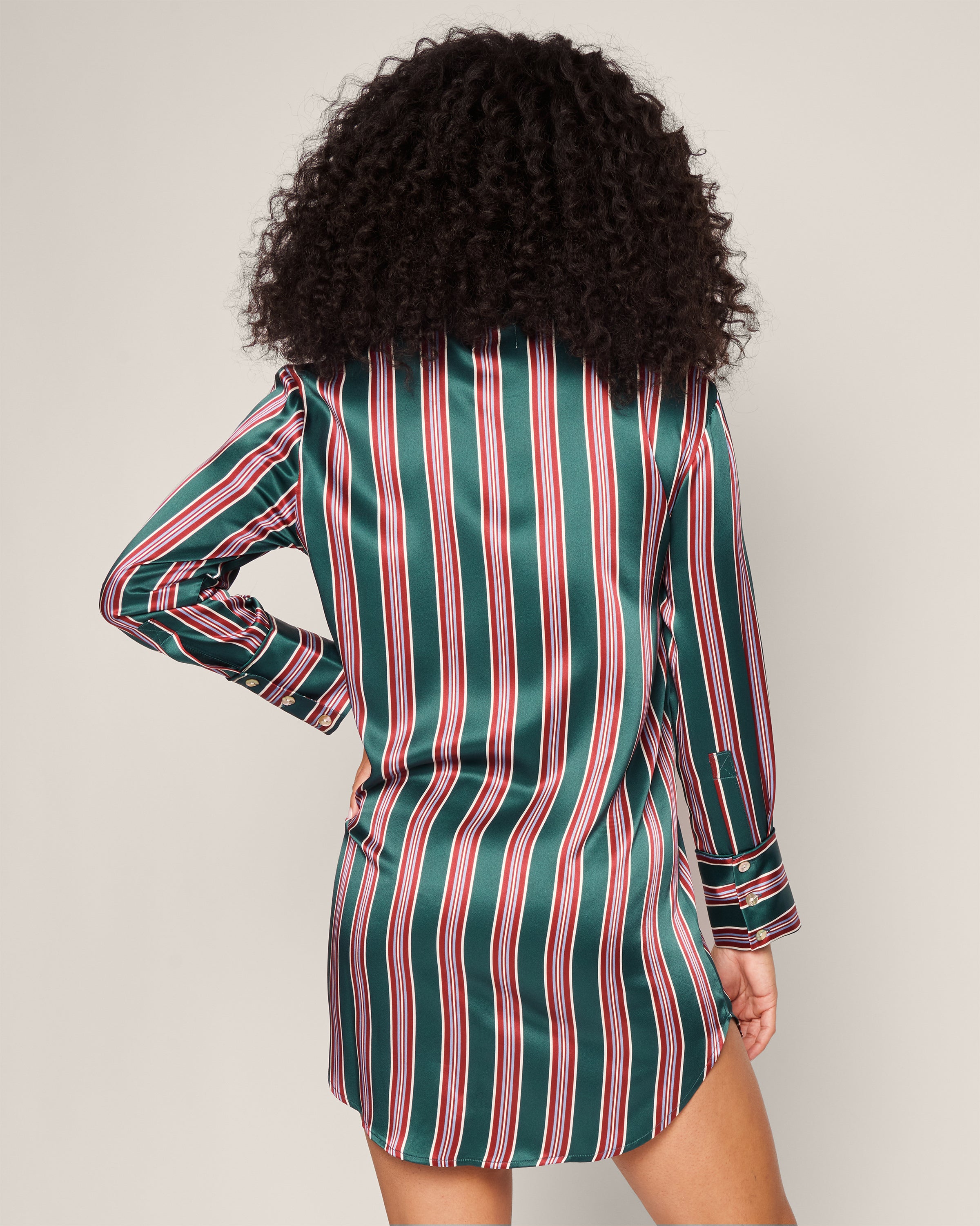 A rear view shows someone with curly hair wearing a Womens Silk Nightshirt in Lancaster Stripe by Petite Plume. This long-sleeve, striped dress in green, red, and white features vertical stripes and buttoned cuffs. It exudes chic pajama vibes against the plain beige background.