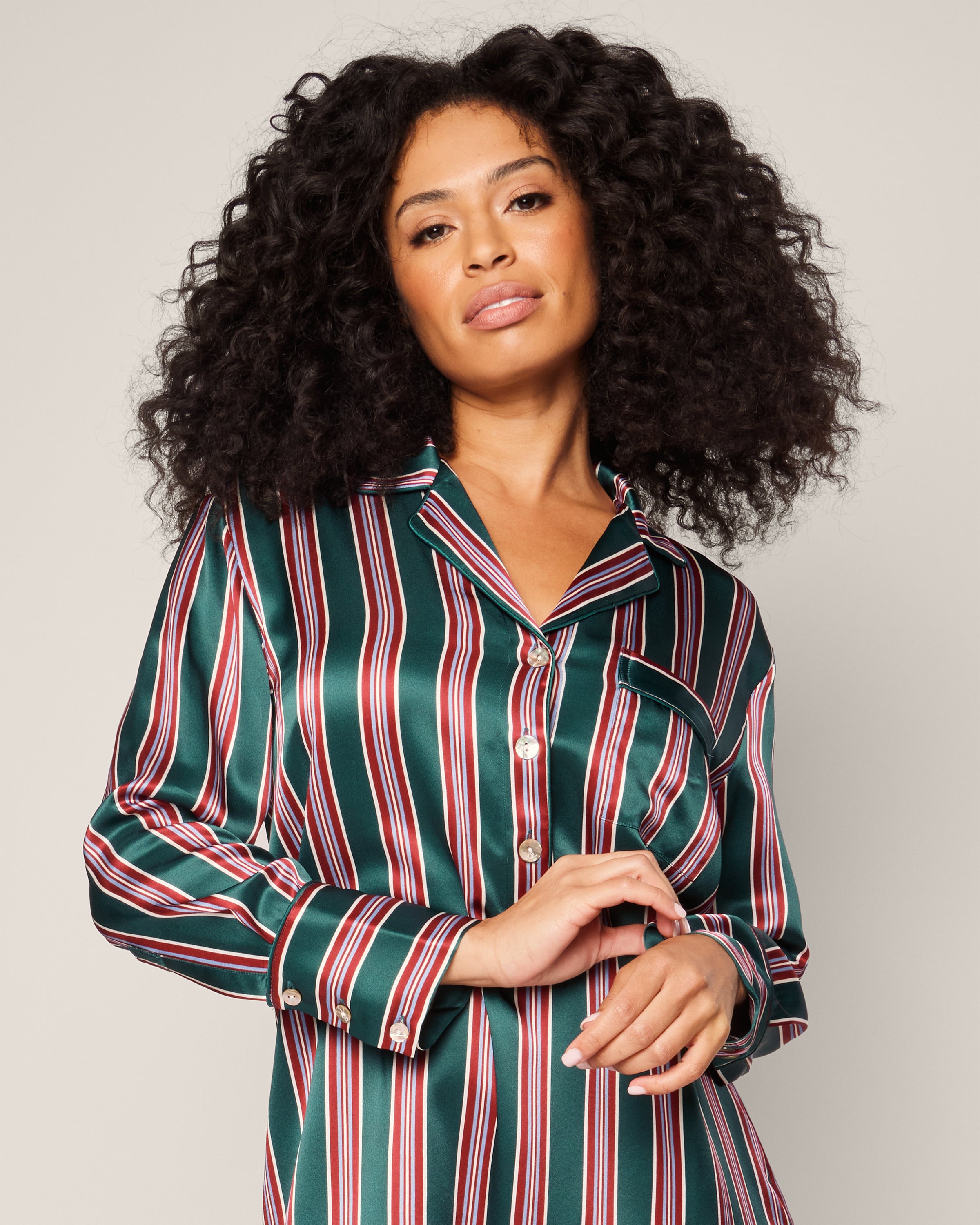 A woman with voluminous curly hair stands confidently against a neutral background, wearing Petite Plumes Womens Silk Nightshirt in Lancaster Stripe, crafted from 22mm Mulberry Silk, her hands gently clasped in front, exuding the elegance of luxurious sleepwear.