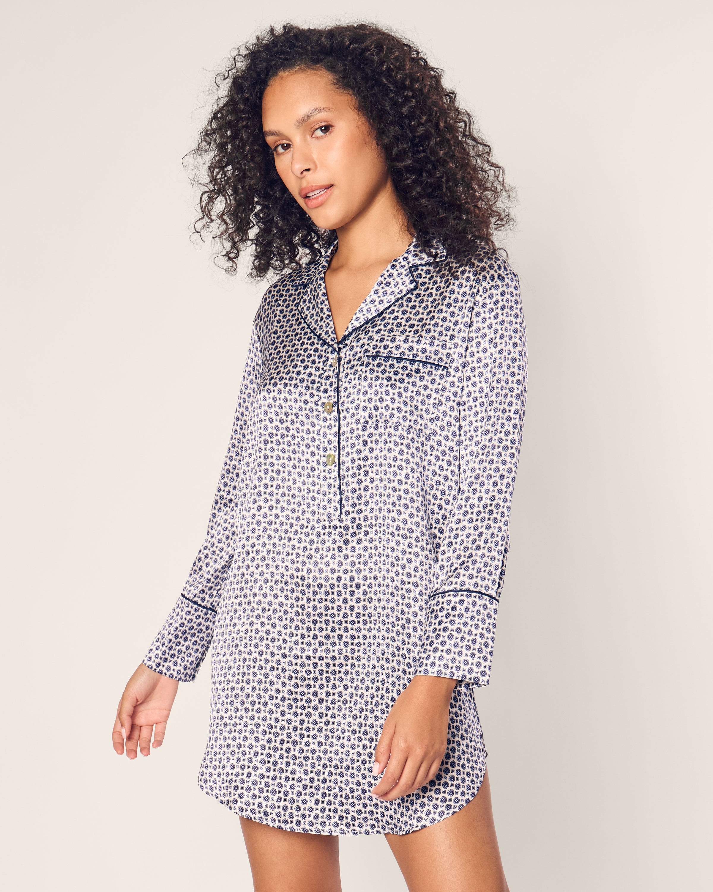 A woman with curly hair stands against a light background, wearing a Petite Plume Womens Silk Nightshirt in Navy Foulard, gazing slightly to the side.