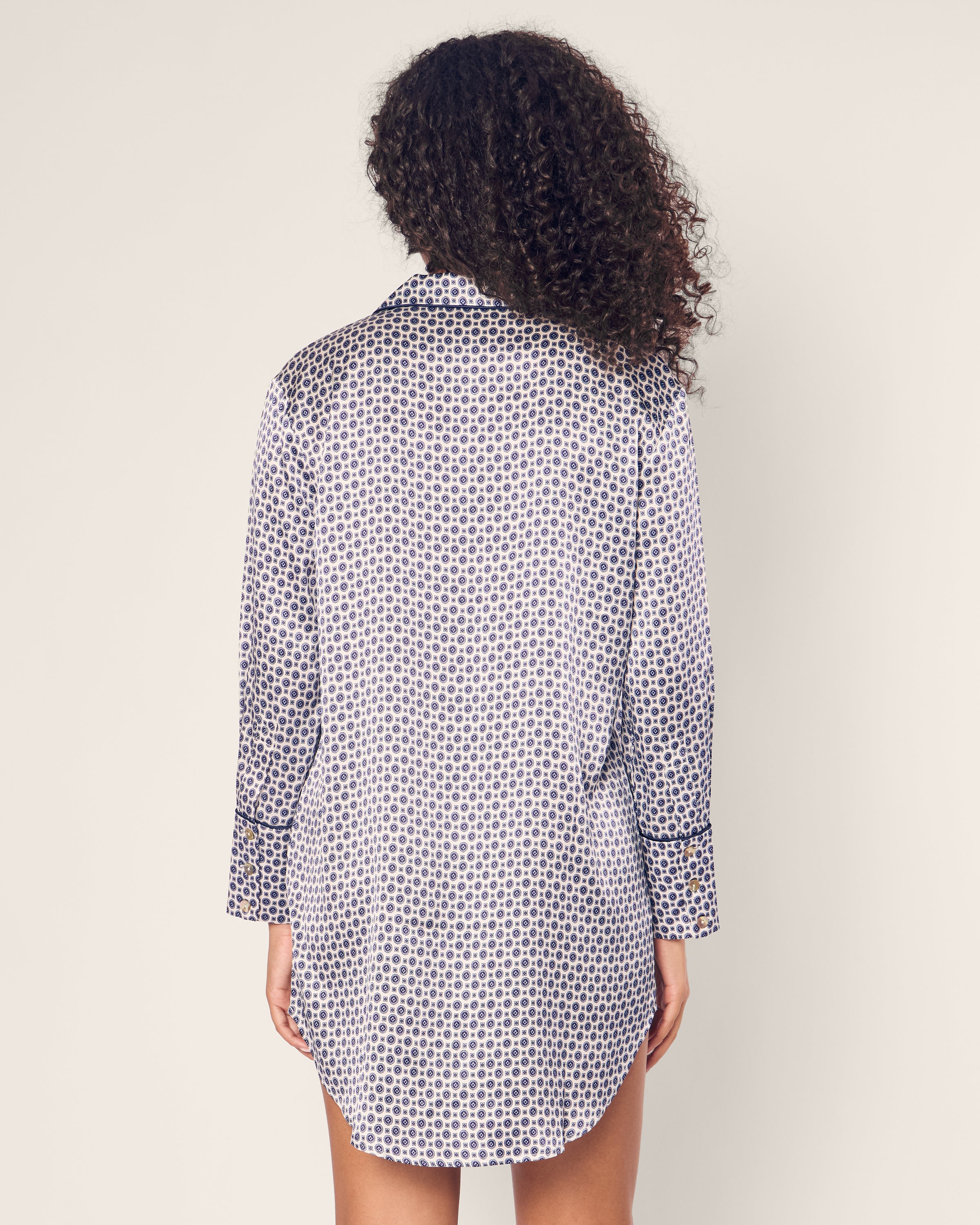 A person with curly hair is seen from the rear wearing Petite Plume’s Womens Silk Nightshirt in Navy Foulard. Crafted from Mulberry silk, the long-sleeve shirt features a black and white circular design with cuff and collar trim, exuding elegant sleepwear vibes against a plain light background.