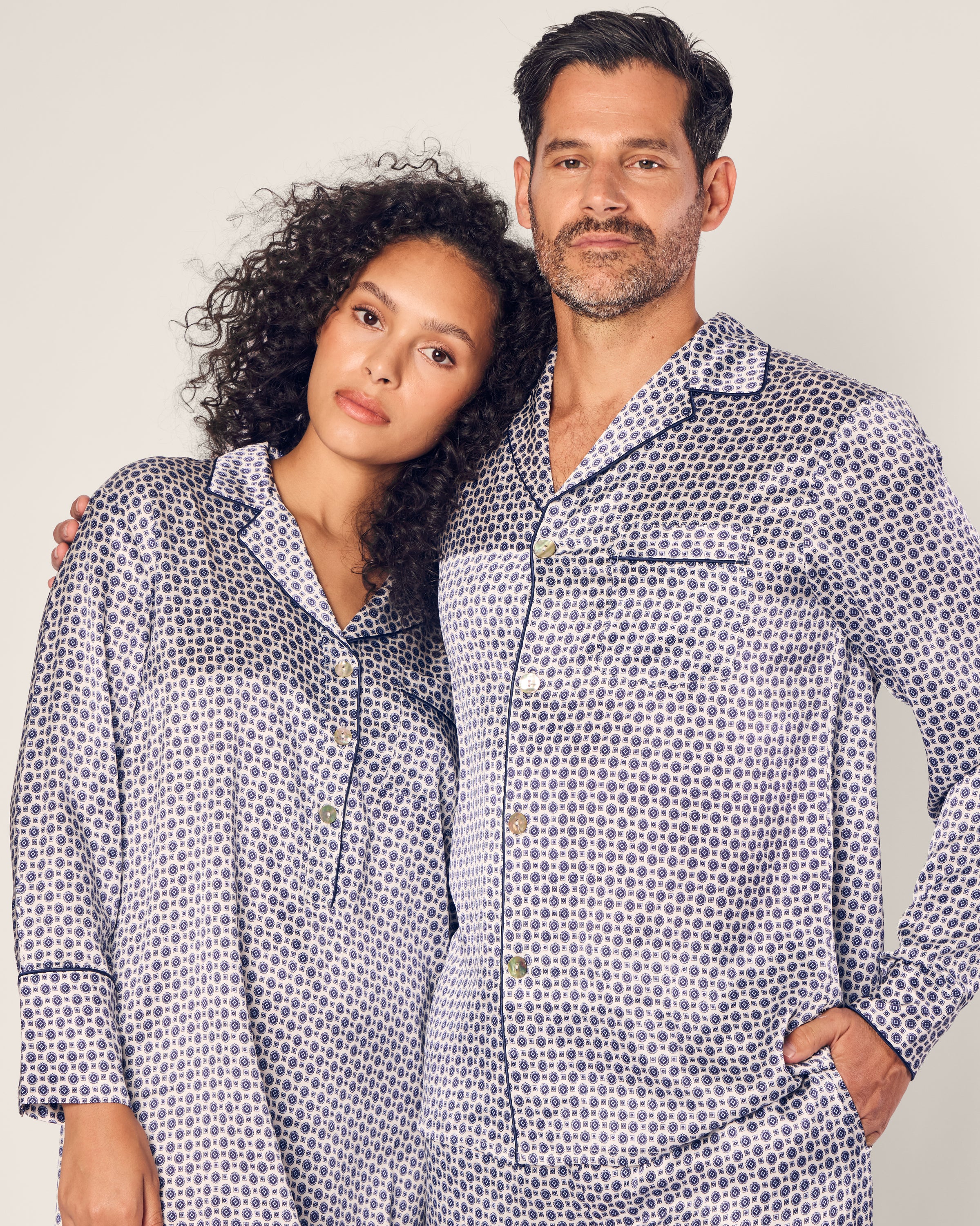 A man and a woman wear matching pajamas; hers is a Womens Silk Nightshirt in Navy Foulard by Petite Plume. They stand close together, their sleepwear showcasing dark circular patterns and black piping on a white backdrop.