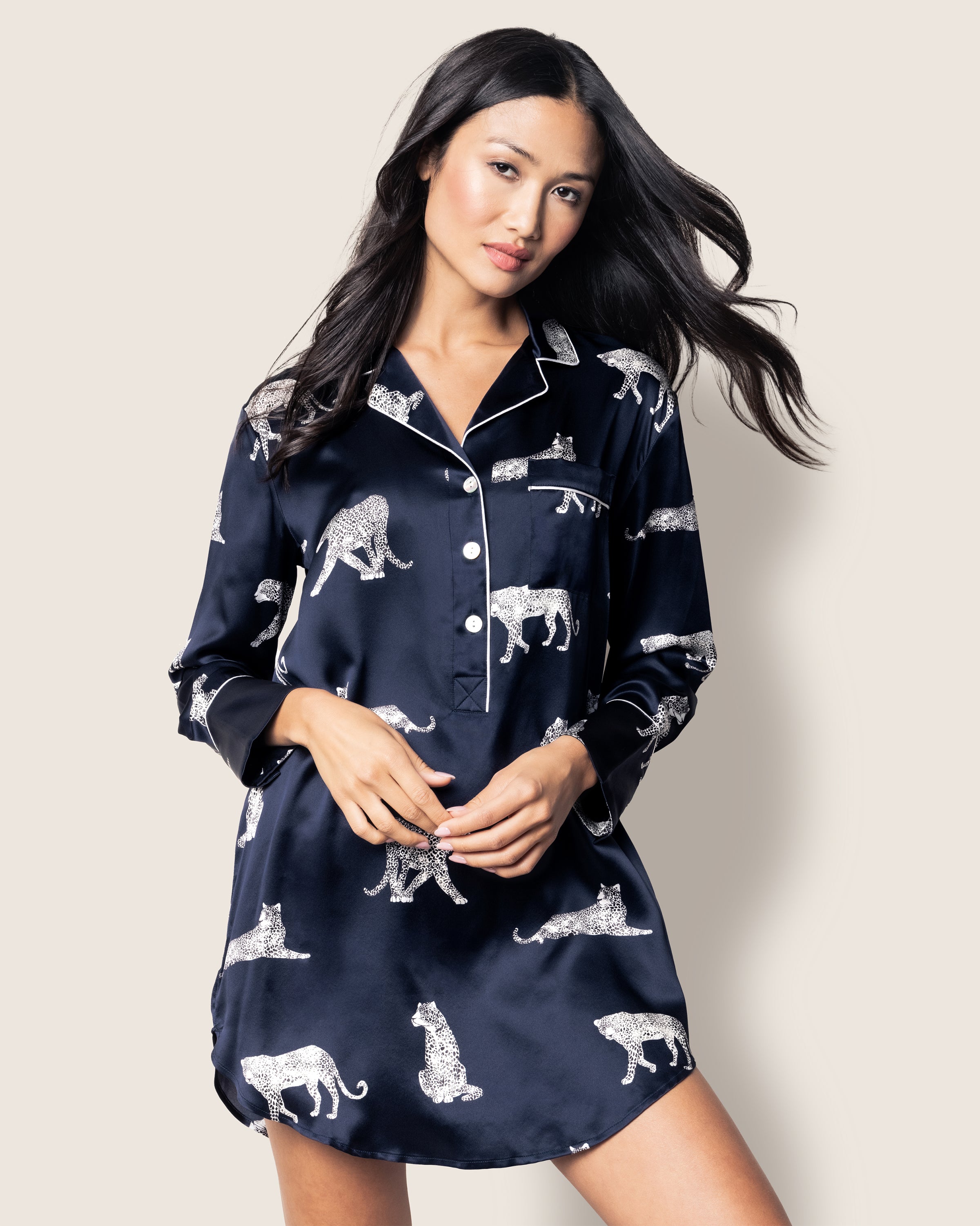 Wearing Petite Plumes Womens Silk Nightshirt in Panthère de Nuit, a woman showcases a navy blue dress adorned with white animal prints like leopards and zebras. Its button-down front and collar evoke elegant sleepwear. Her long, dark hair contrasts with the light beige background.