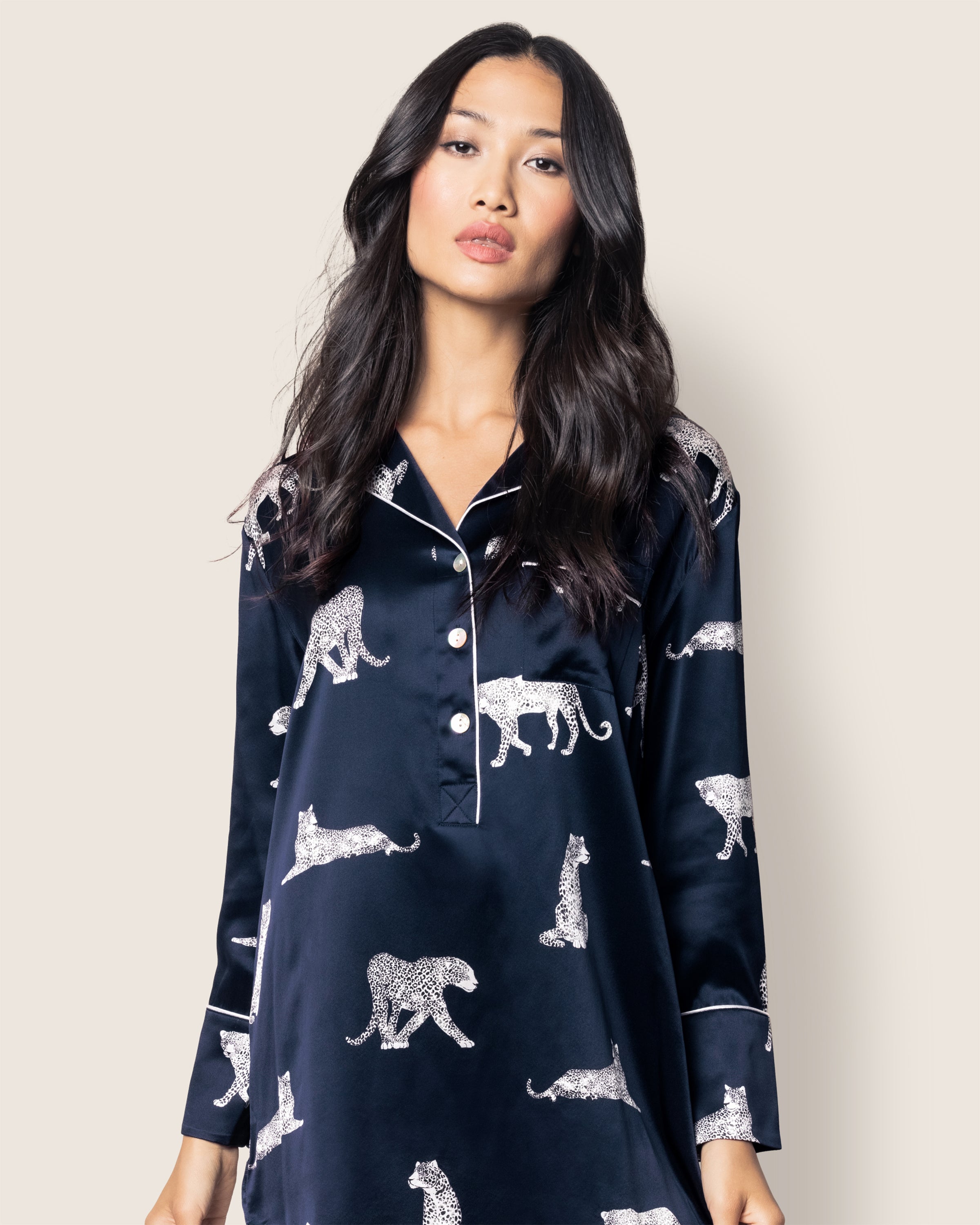 A person with long black hair wears Petite Plumes Womens Silk Nightshirt in Panthère de Nuit, crafted from dark Mulberry silk with a white leopard pattern and a light plain background.