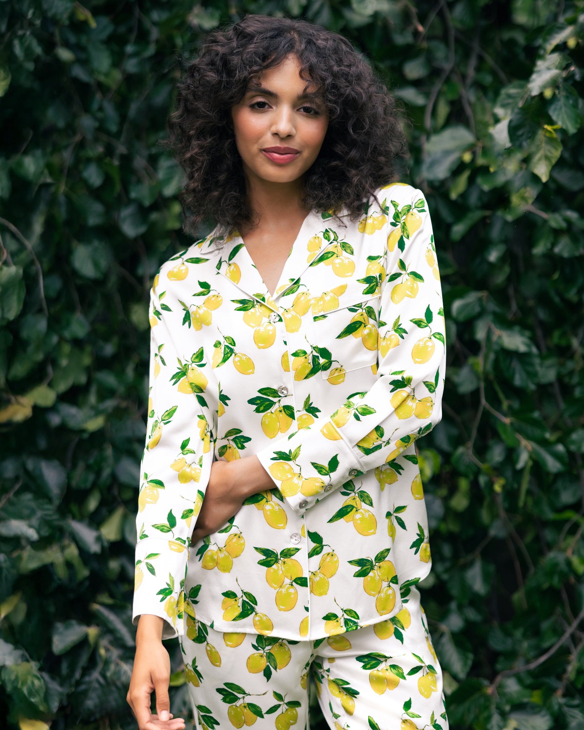 A woman with curly hair wearing Petite Plumes Womens Silk Pajama Set in Amalfi Lemons stands smiling slightly against lush greenery. The limited-edition silk set adds luxury to her vibrant look.