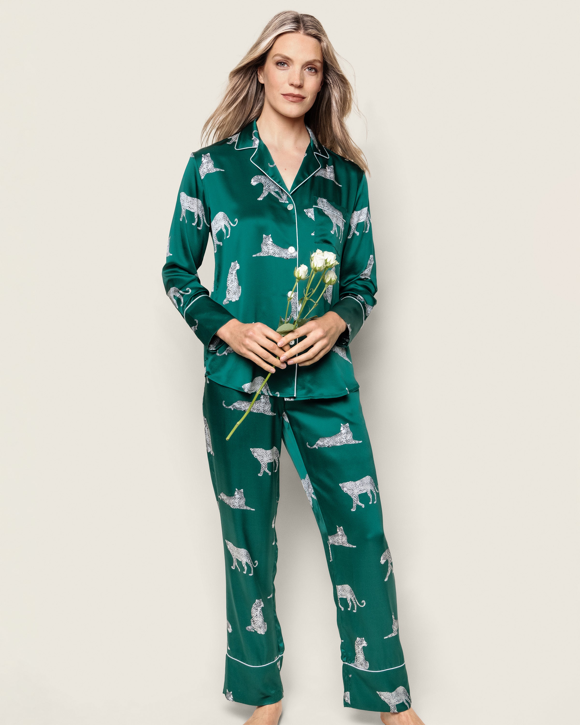 A person with long hair stands against a plain background, wearing the Petite Plume Womens Silk Pajama Set in Panthère de Luxe and holding white flowers, exuding a relaxed expression.