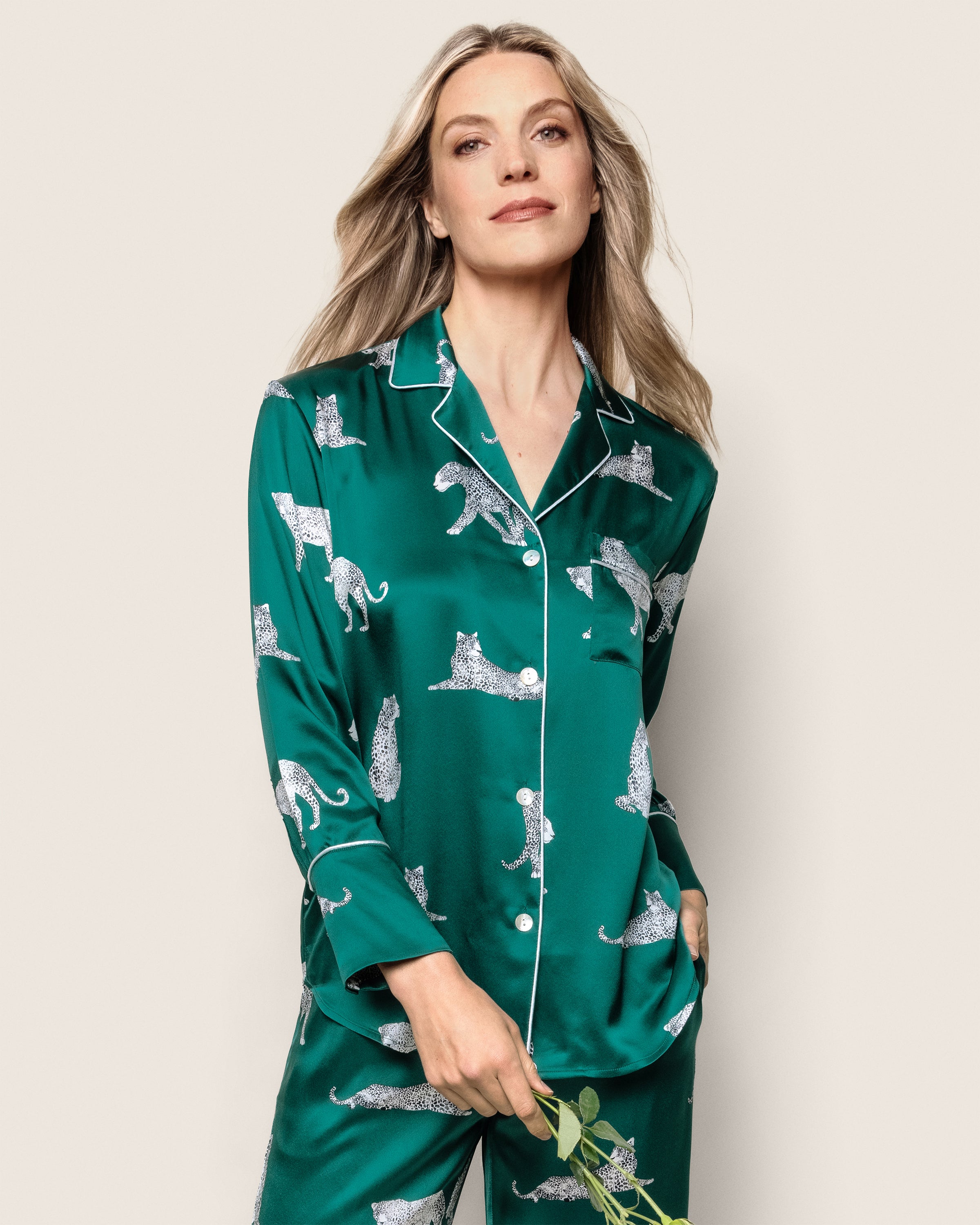 A person with long blonde hair is wearing a luxurious Petite Plume Womens Silk Pajama Set in Panthère de Luxe. They are holding a small branch with green leaves and standing against a plain background.