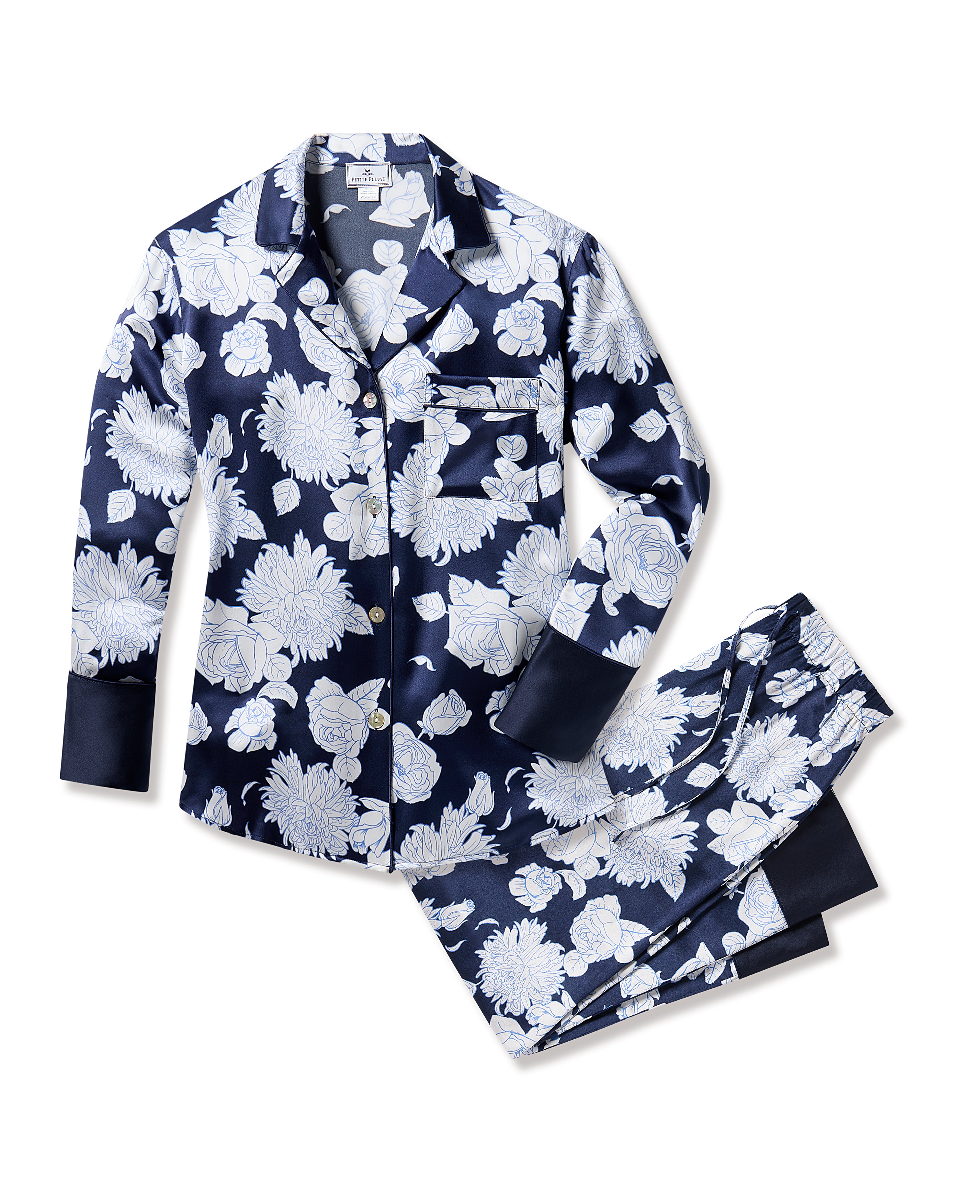 Treat yourself to the luxury of Petite Plumes Womens Silk Pajama Set in Midnight Blooms. Made from machine-washable silk, this navy set features a white floral pattern. The top has long sleeves, a collar, front buttons, and a chest pocket. The pants offer an elastic waistband with dark cuffs.