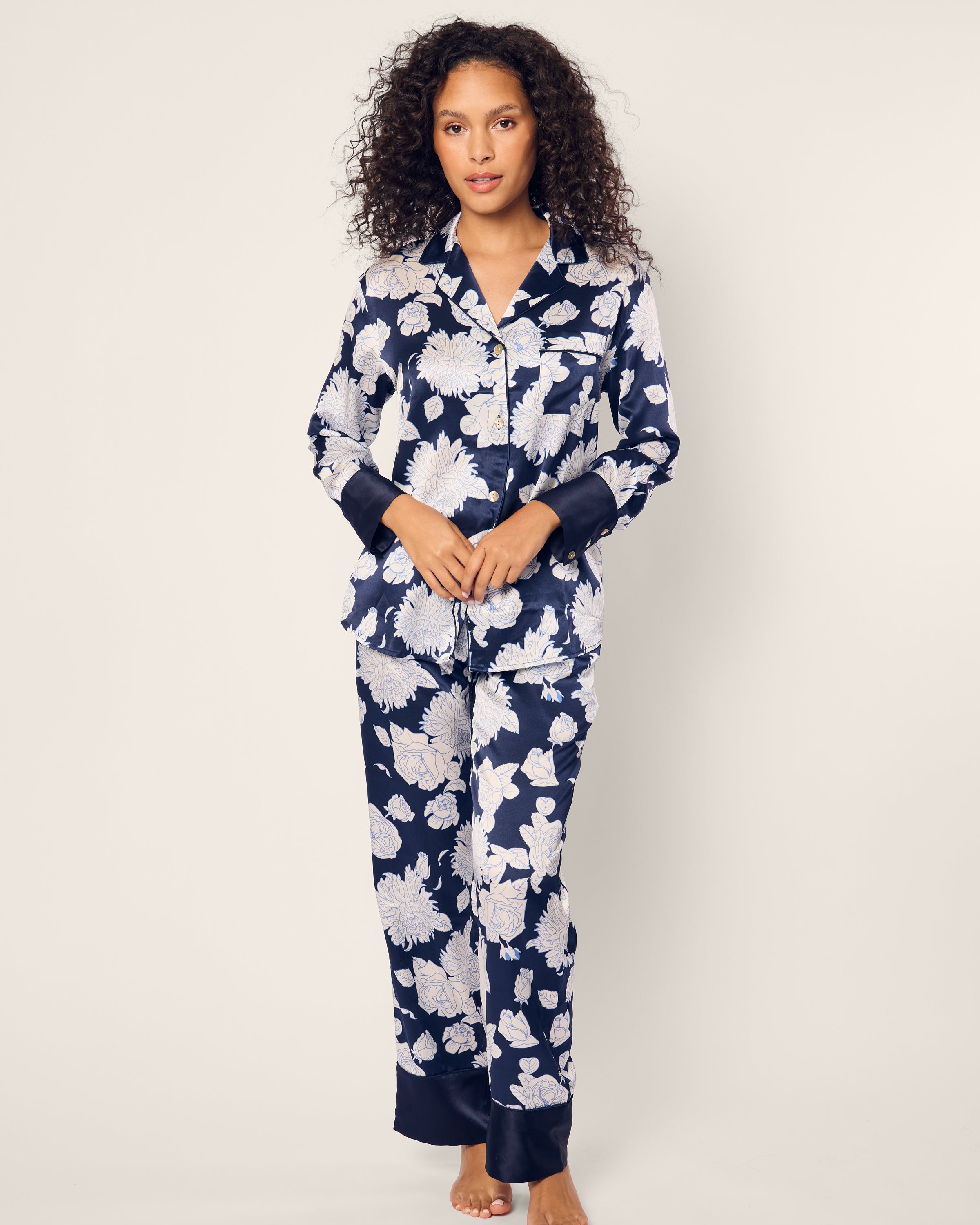 A woman with curly hair models the Womens Silk Pajama Set in Midnight Blooms by Petite Plume, featuring luxurious navy mulberry silk with large white florals. She stands against a plain light background, gazing directly at the camera with a neutral expression.