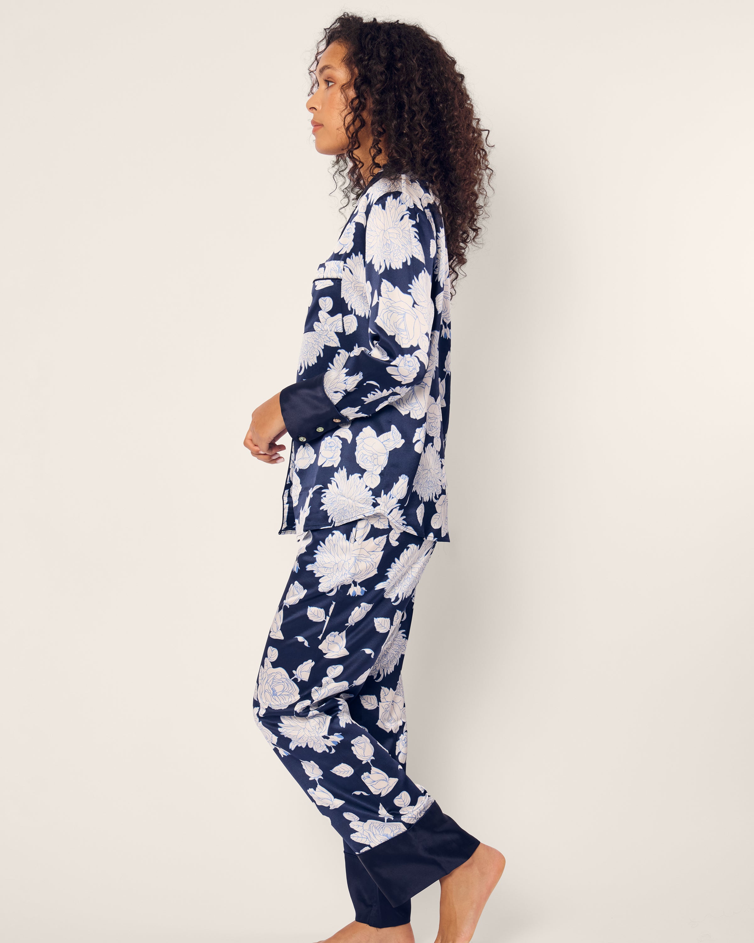 A person with curly hair stands sideways, showcasing Petite Plumes luxurious Womens Silk Pajama Set in Midnight Blooms. Made from rich mulberry silk, this navy set with a white floral pattern exudes elegance while being machine-washable. They are barefoot against a plain light background.