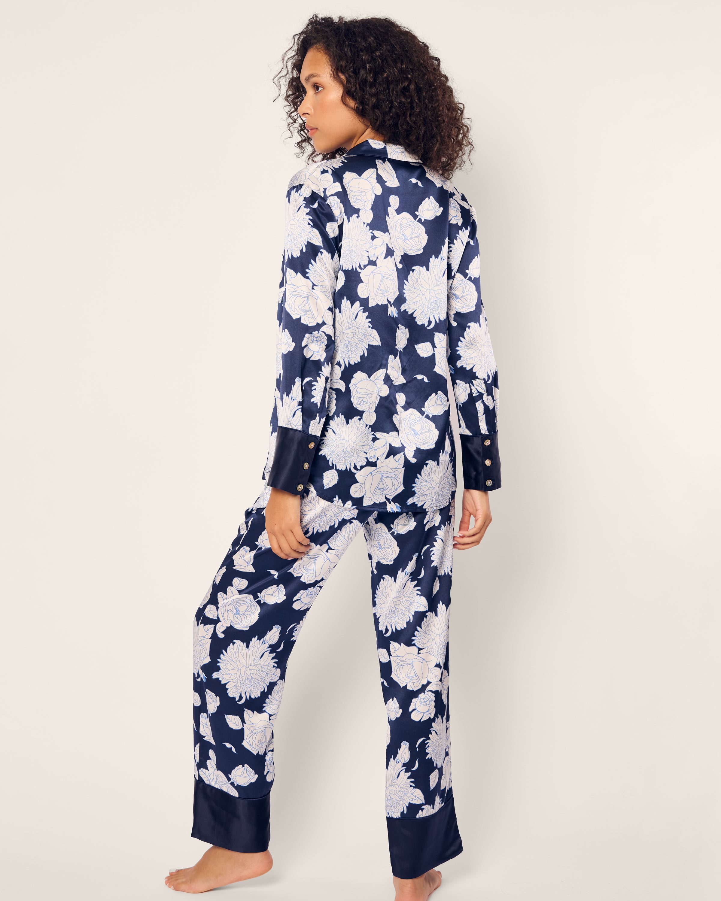 A person with curly hair wears the Womens Silk Pajama Set in Midnight Blooms by Petite Plume. The navy blue set features luxurious mulberry silk with a white floral pattern. Theyre standing slightly turned against a plain background.