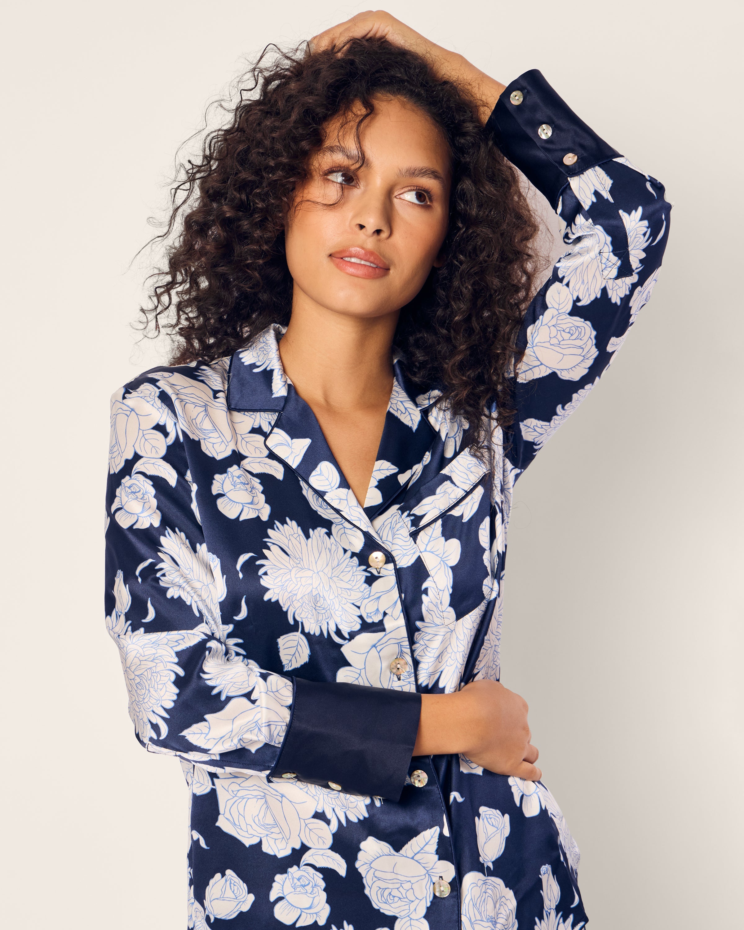 A person with curly hair confidently poses, hand on head and hip, wearing the Womens Silk Pajama Set in Midnight Blooms by Petite Plume. The background is a light neutral color.