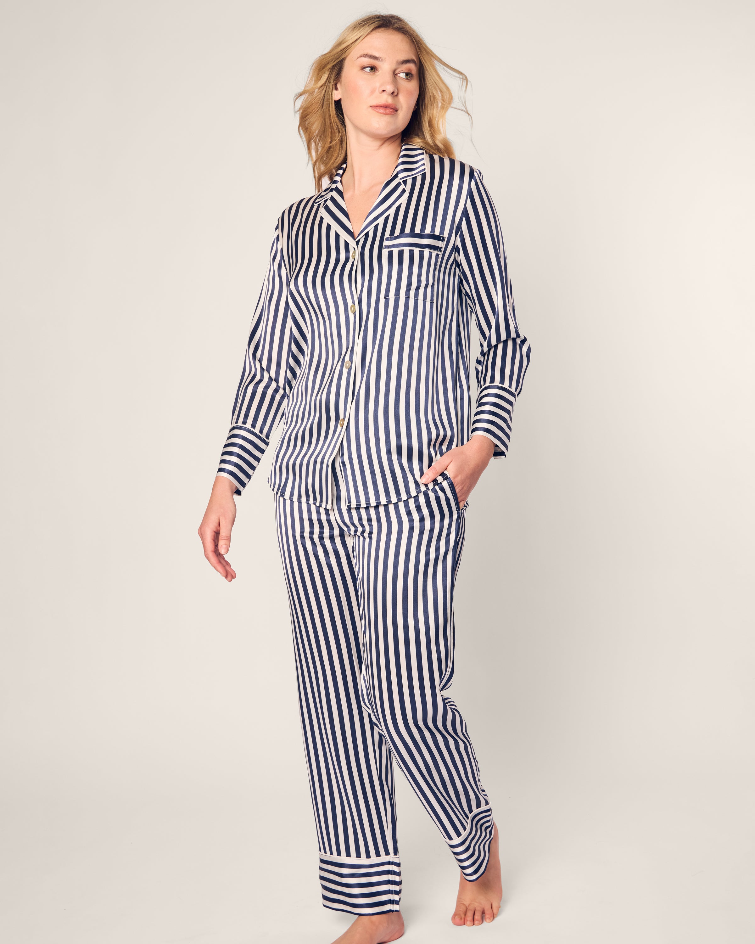 A person exudes effortless elegance in a Womens Silk Pajama Set from Petite Plume, featuring a luxurious navy Bengal stripe with a long-sleeve button-up shirt and pants, as they walk casually with one hand in a pocket against a plain background.