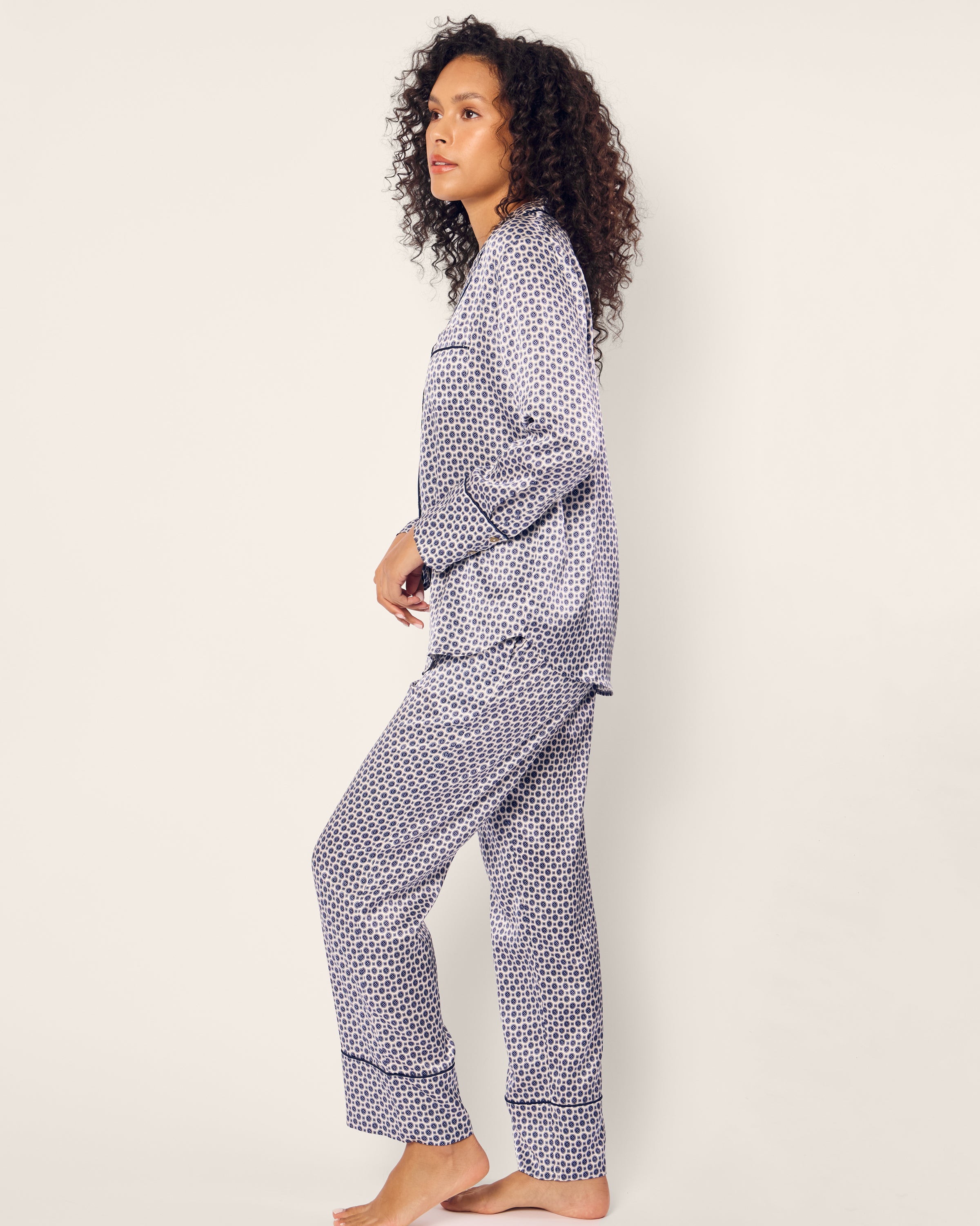 A person with curly hair stands barefoot against a plain background, wrapped in Petite Plumes luxury Womens Silk Pajama Set in Navy Foulard. The patterned, long-sleeved top and pants exude elegance as they face sideways, maintaining a relaxed posture and neutral expression.