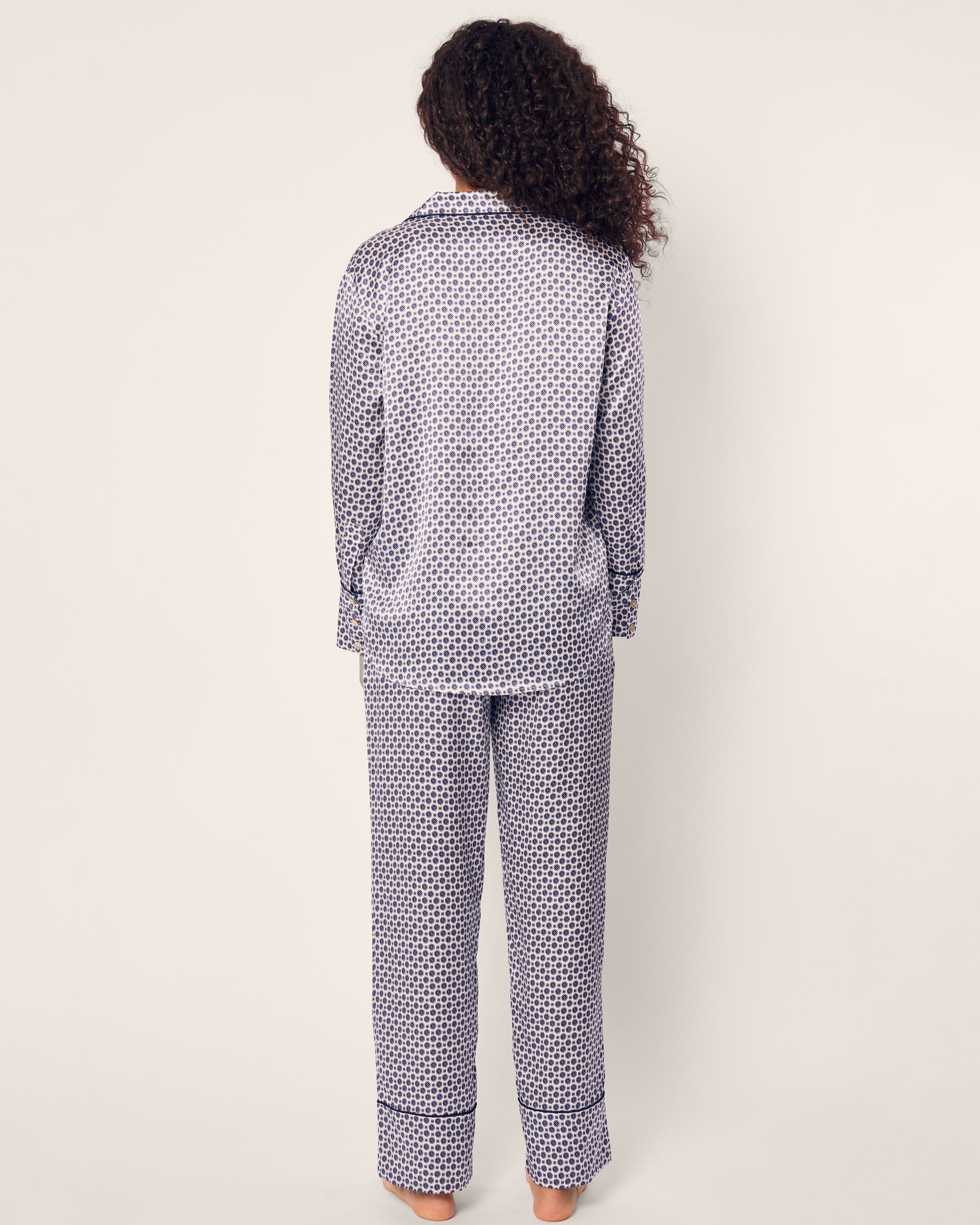 A person with curly hair wears Petite Plumes Womens Silk Pajama Set in Navy Foulard, barefoot against a plain light background, exuding elegance and comfort.