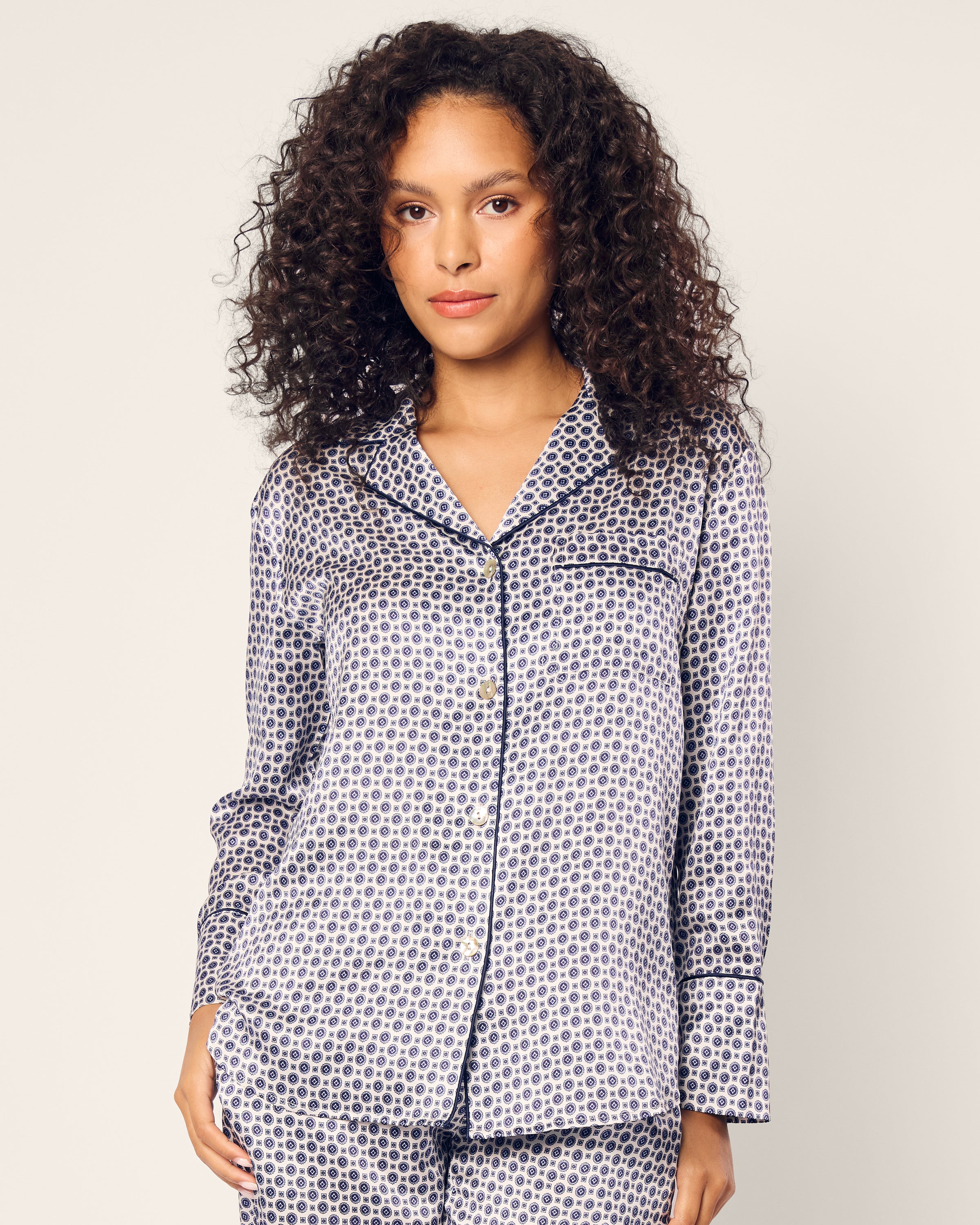 A person with curly hair wears Petite Plumes Womens Silk Pajama Set in Navy Foulard, featuring a dark circular pattern on a light background. They stand against a plain, light backdrop, gazing at the camera, epitomizing luxurious sleepwear.