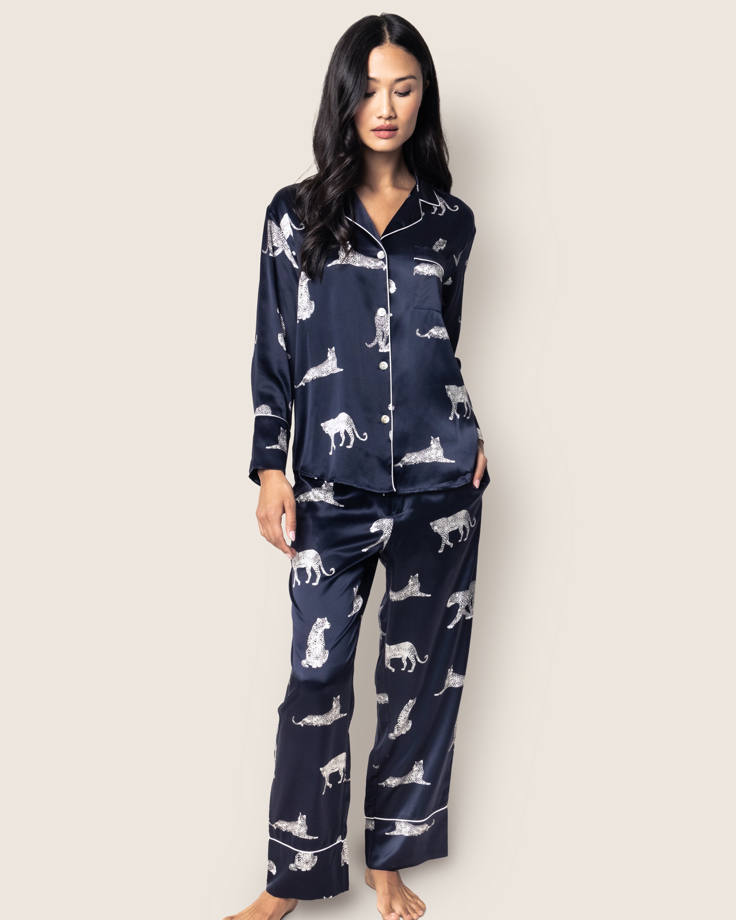 A person with long black hair wears Petite Plumes Womens Silk Pajama Set in Panthère de Nuit, featuring elegant navy blue pajamas adorned with panther prints. Against a plain backdrop, they confidently pose with one hand in the pocket, embodying luxury sleepwear charm.