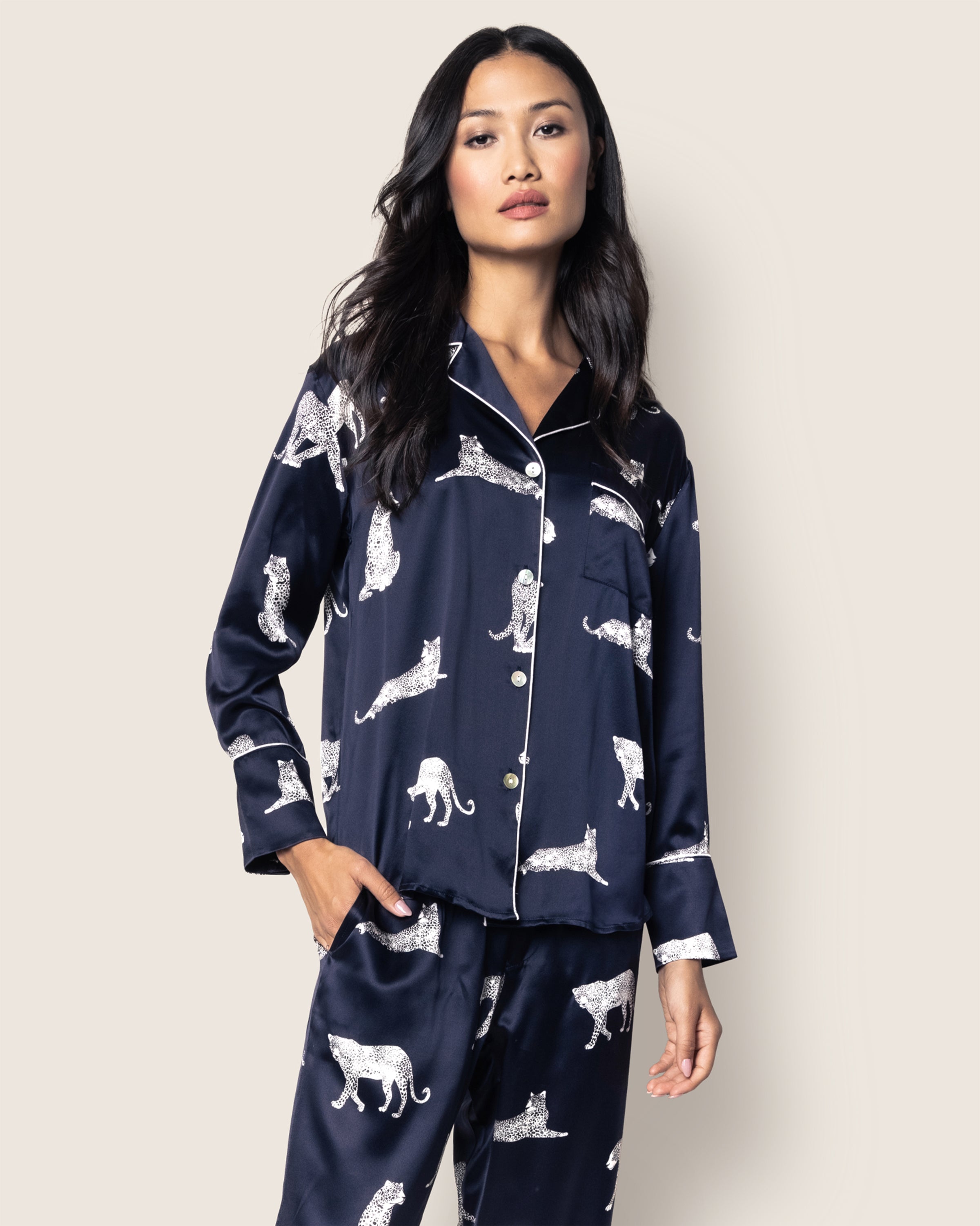 A person models Petite Plumes Womens Silk Pajama Set in Panthère de Nuit against a light backdrop. The navy set with elegant panther prints features a buttoned top and collar, while their long dark hair drapes gracefully over their shoulders and their hands rest relaxed at their sides.