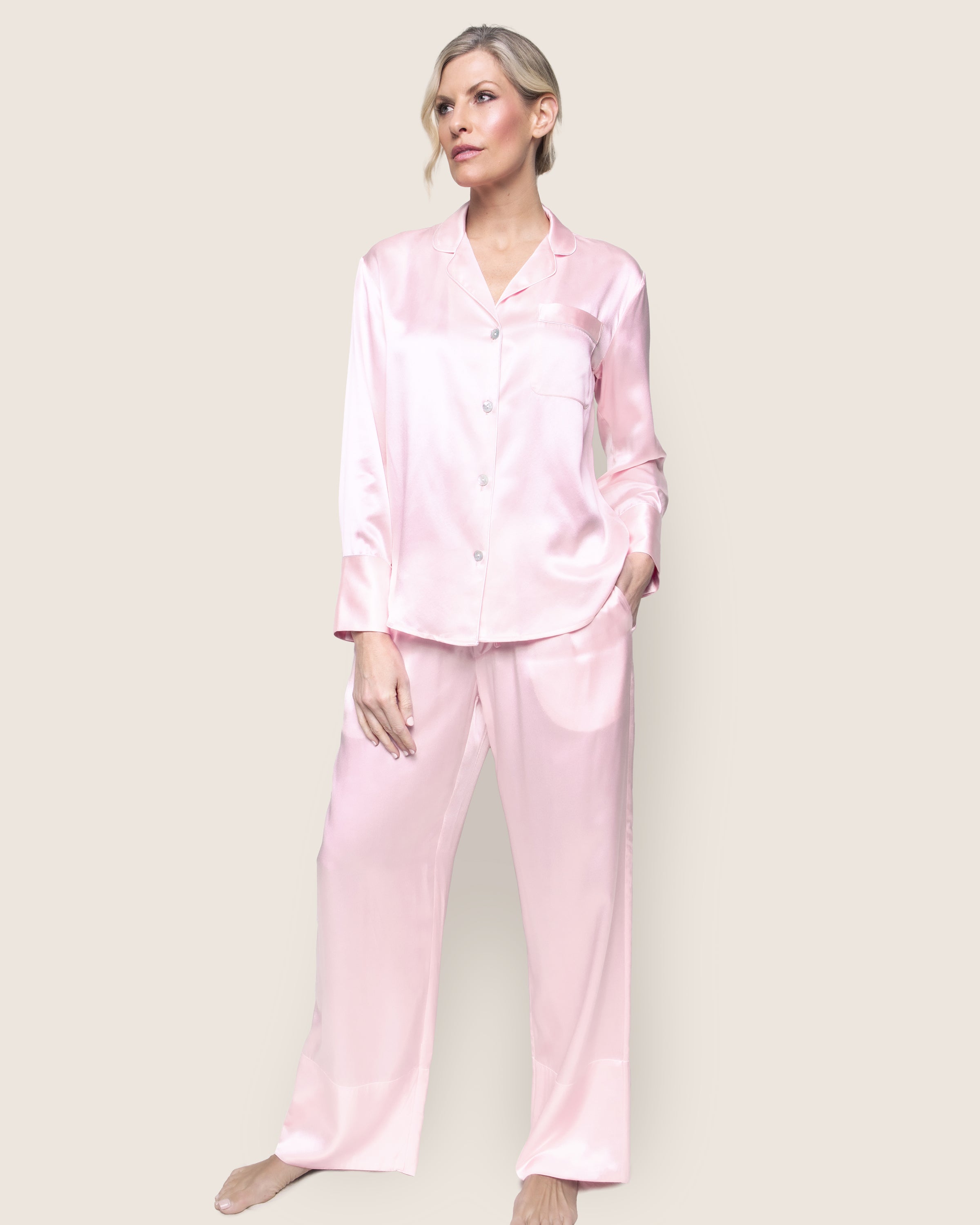 Wearing the Womens Silk Pajama Set in Pink by Petite Plume, a person poses against a light background. This luxurious sleepwear includes a button-up top with a pocket and long pants, offering an elegant and comfortable look.