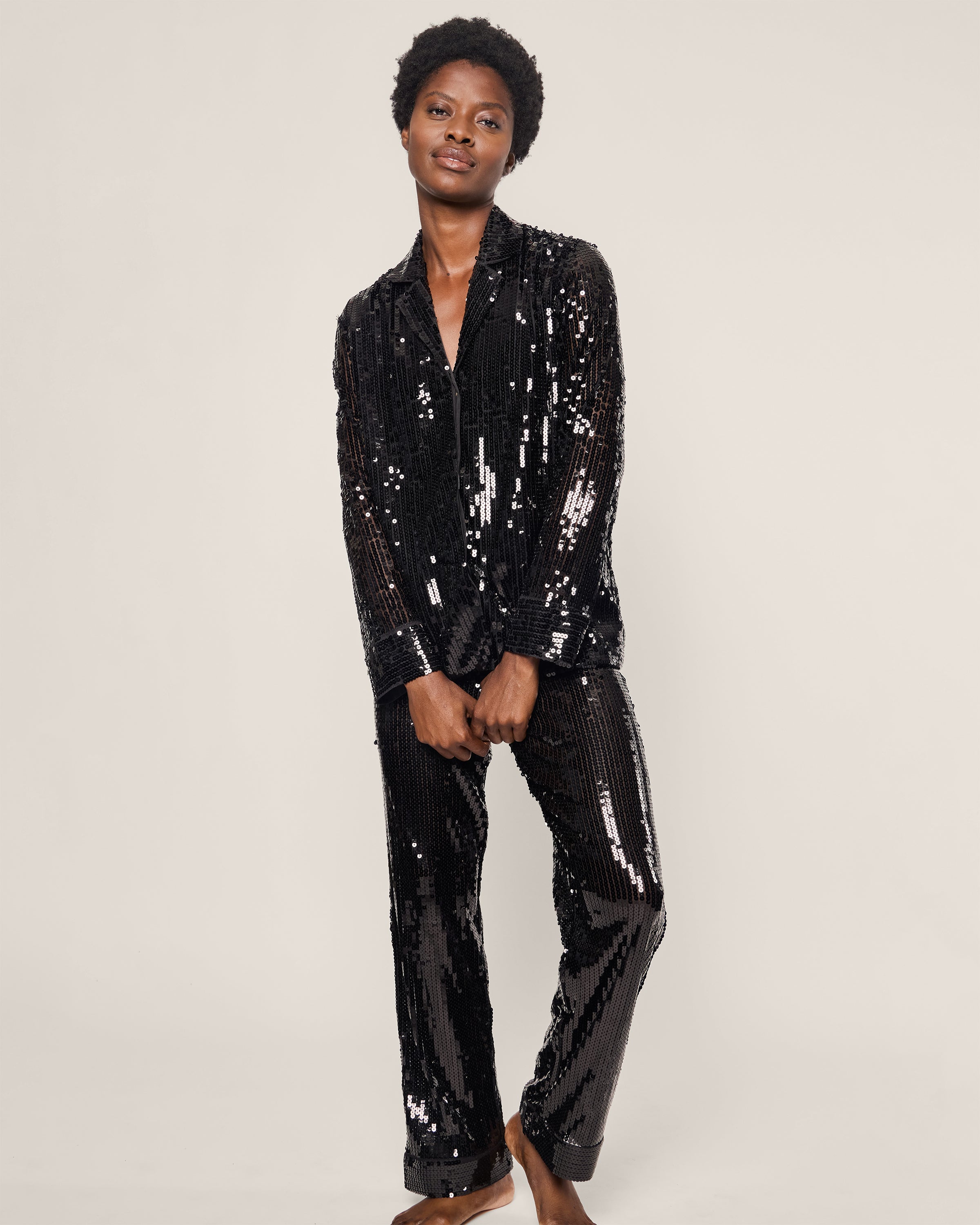 Clad in a Petite Plume Womens Pajama Set in Black Sequins, a person stands barefoot with a slight smile. Their relaxed arms and the elegant pajama silhouette exude luxury, set against a plain background.