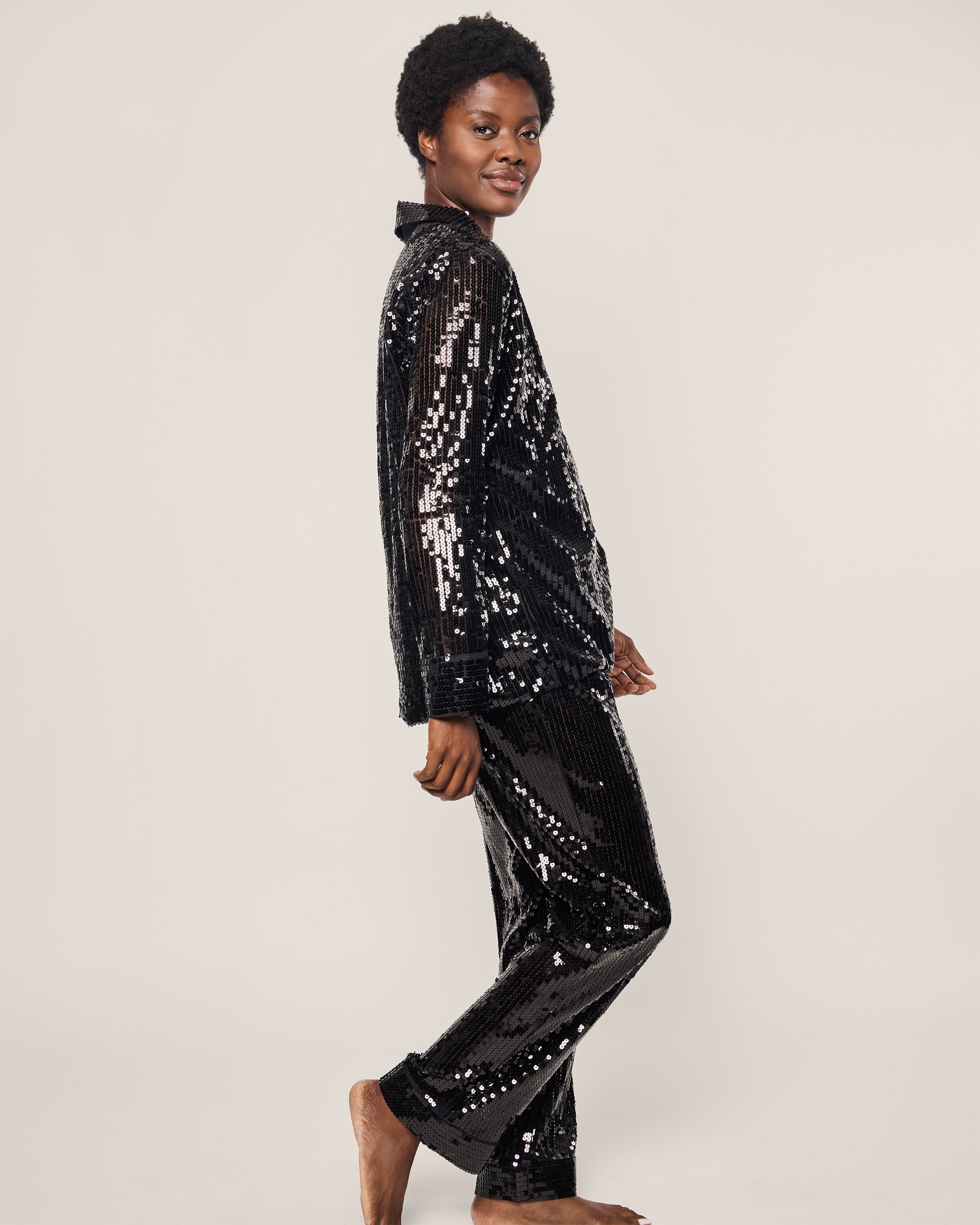 A person smiles in profile, showcasing the Petite Plume Womens Pajama Set in Black Sequins, featuring long sleeves and pants that mirror a classic pajama silhouette. They stand barefoot against a plain light background.