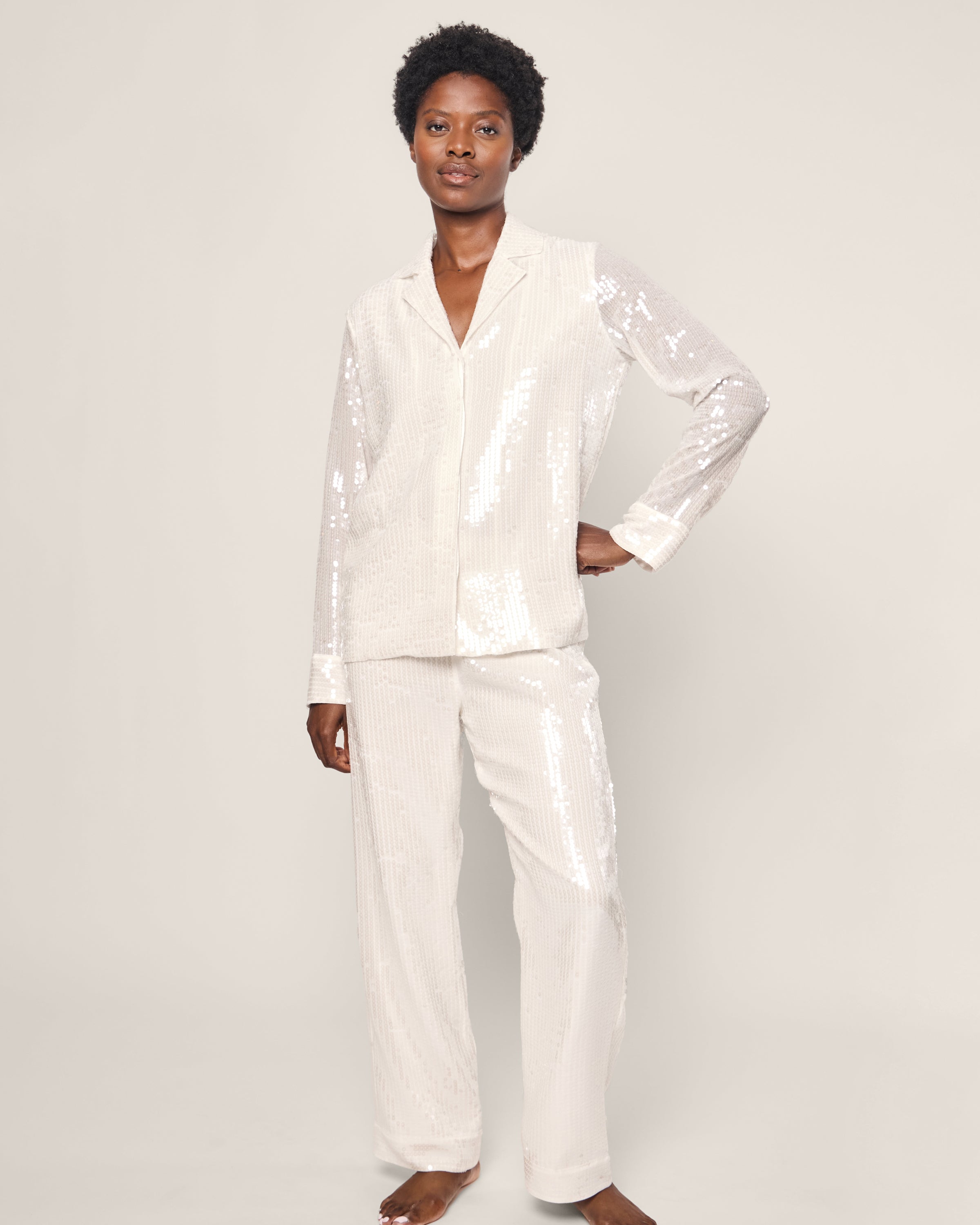 A person models the Petite Plume Womens Sequin Pajama Set in White against a plain backdrop. The classic long-sleeve top and pants shimmer luxuriously, with their hands relaxed by their sides.