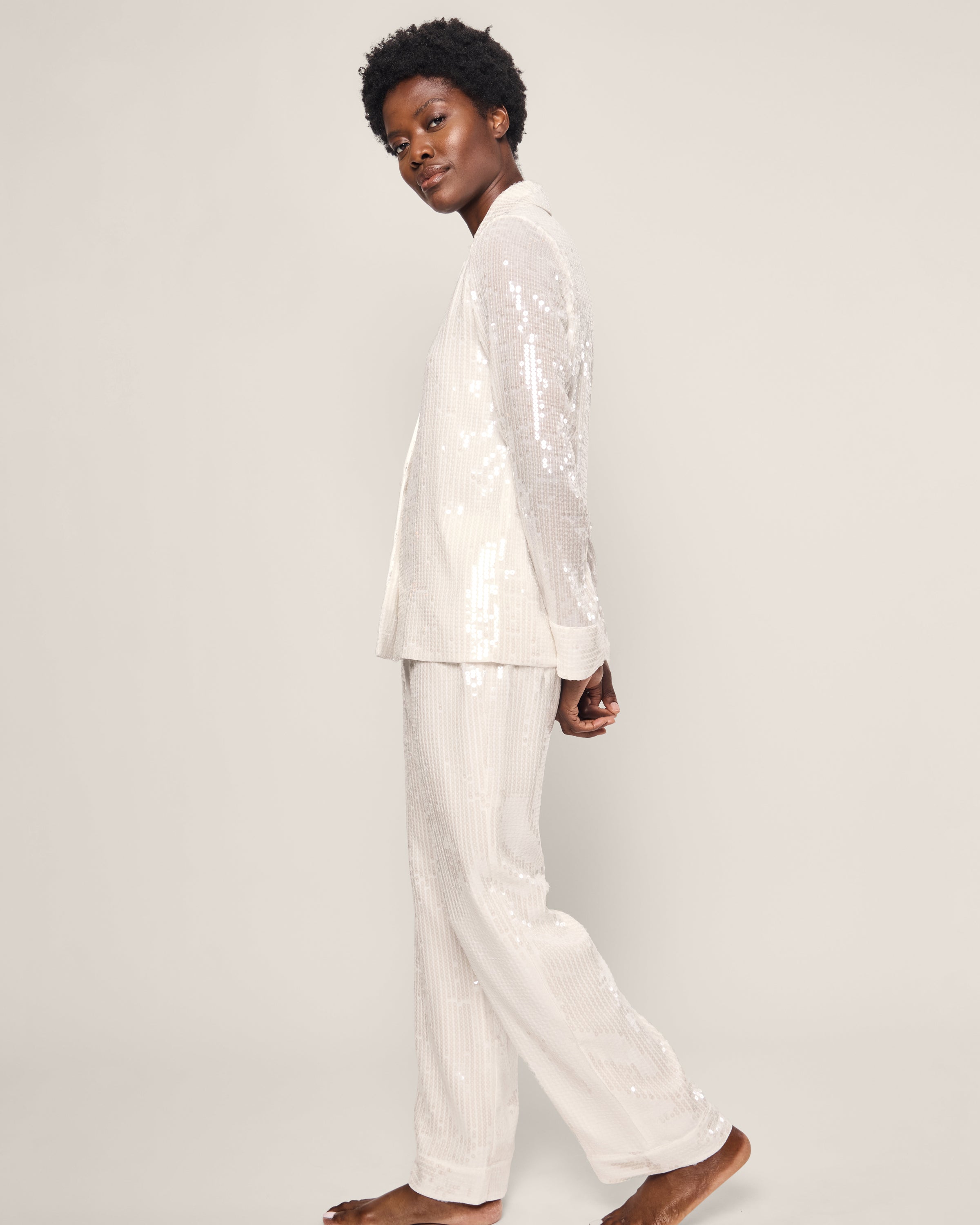 A person wearing the Womens Sequin Pajama Set in White by Petite Plume stands sideways, looking towards the camera. The outfit, with its classic pajama silhouette, includes a long-sleeved jacket and matching pants against a plain, light neutral background.