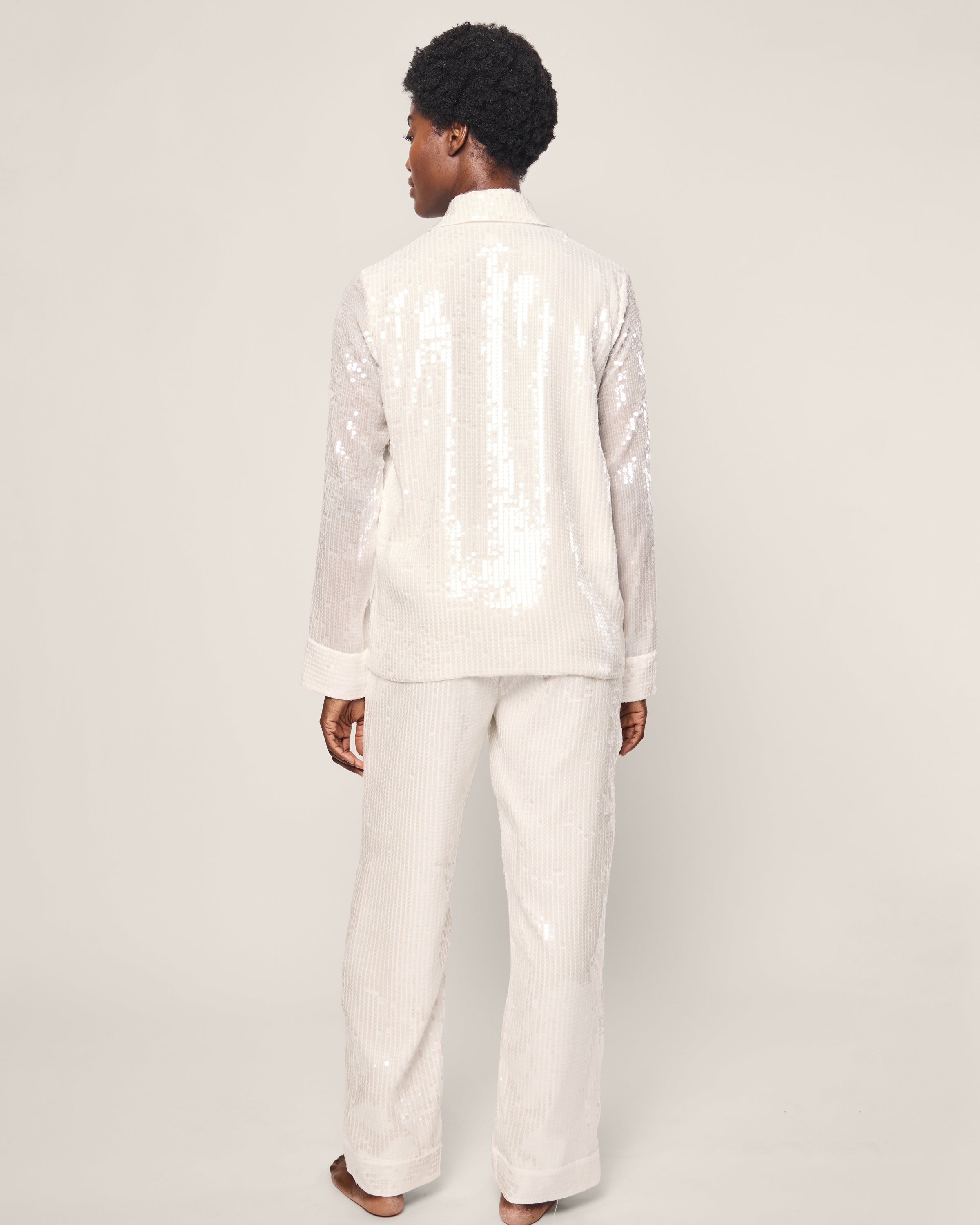 A barefoot model displays the Womens Sequin Pajama Set in White by Petite Plume, featuring a luxurious long-sleeve top and matching pants with white sequins. The classic silhouette and neutral backdrop emphasize the elegance of this sleepwear ensemble.