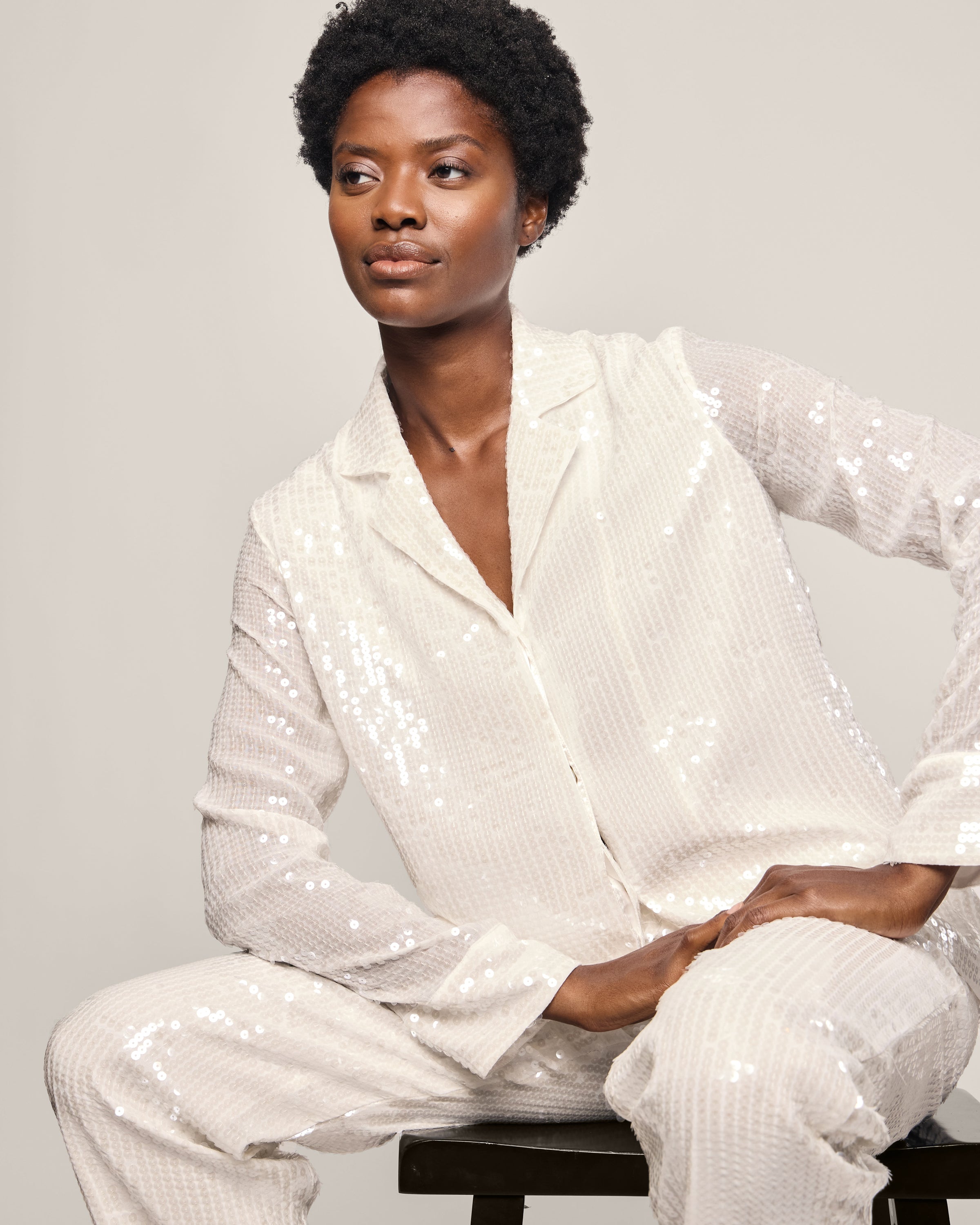 A person with short curly hair poses confidently in a sparkling white Womens Sequin Pajama Set by Petite Plume, set against a plain, light background.