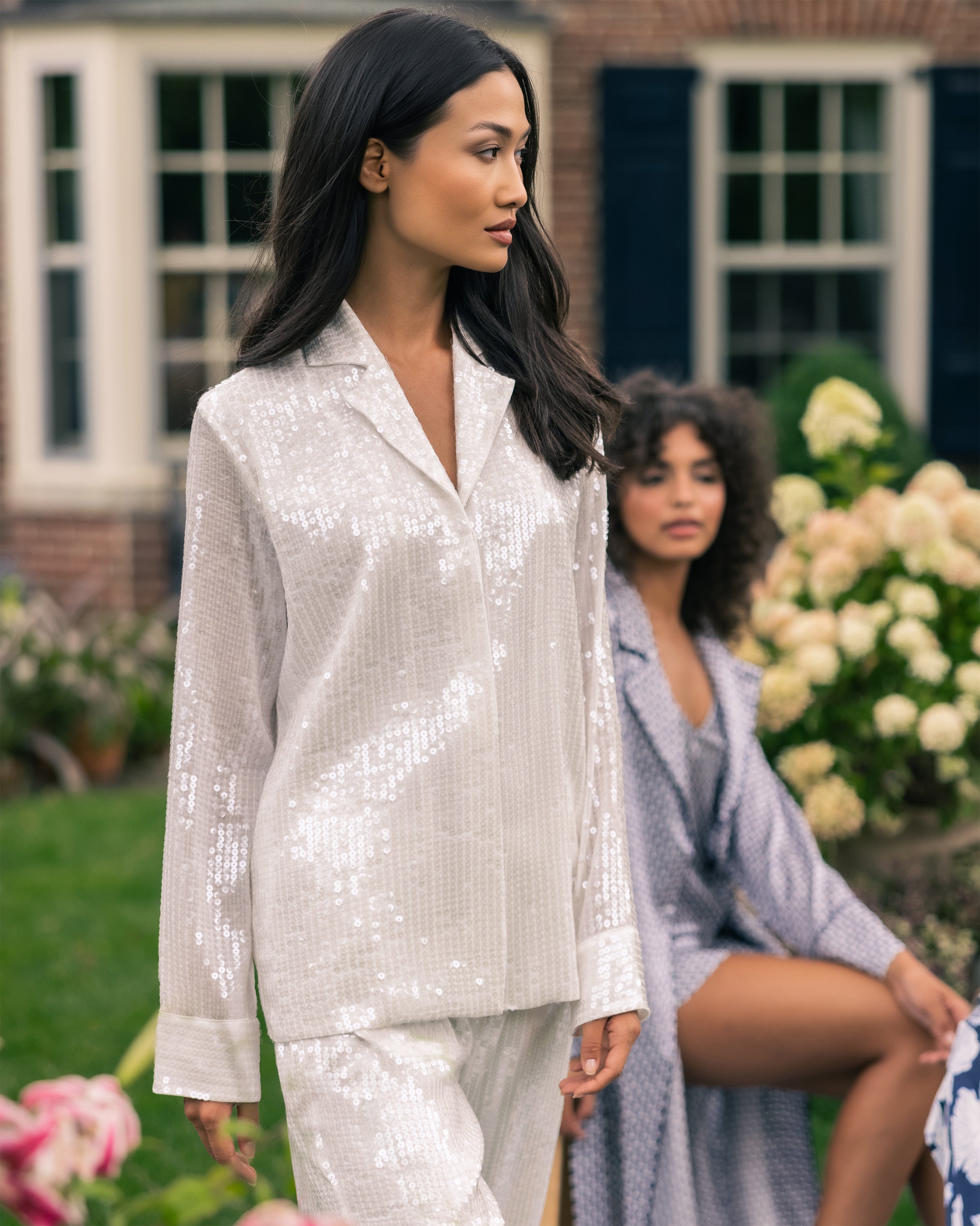 A woman in a Womens Sequin Pajama Set by Petite Plume stands outside, gazing to the side, while another in classic pajamas sits on a bench amid flowers. A charming brick house completes the serene garden scene.